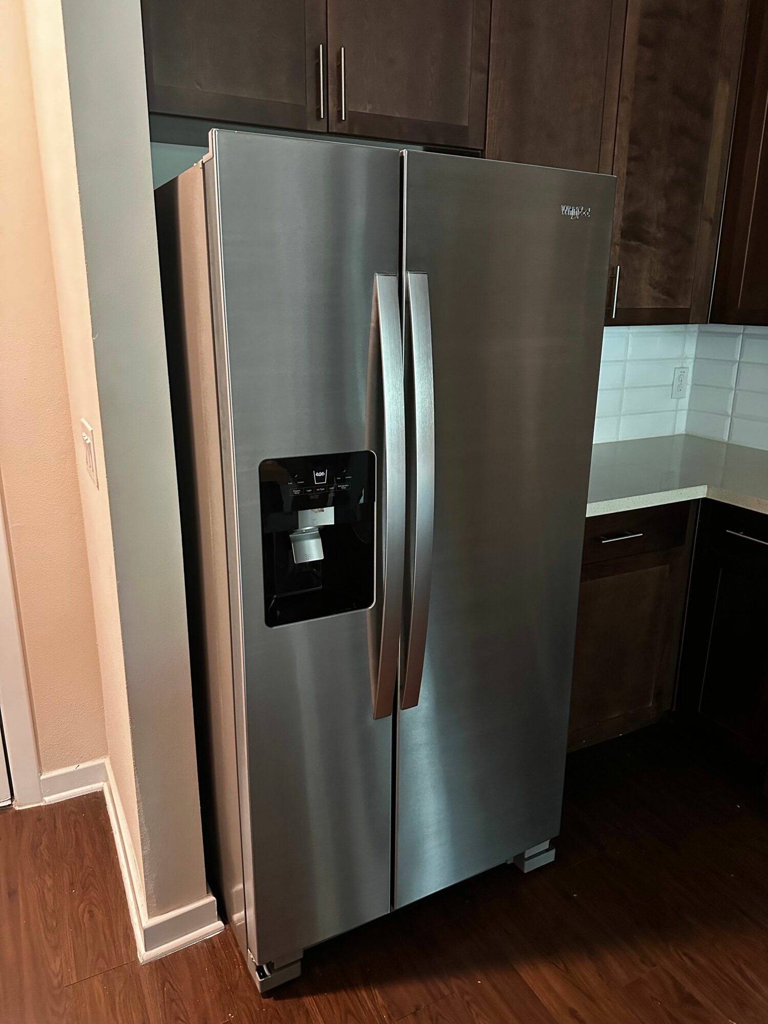 Stainless steel refrigerator with a black control panel, positioned in a kitchen with dark wood flooring and cabinets.