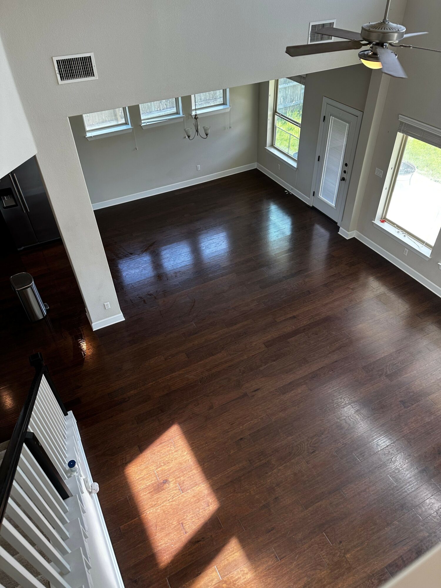 Empty room with hardwood floor, large windows, and a ceiling fan, sunlight casting shadows on the floor.