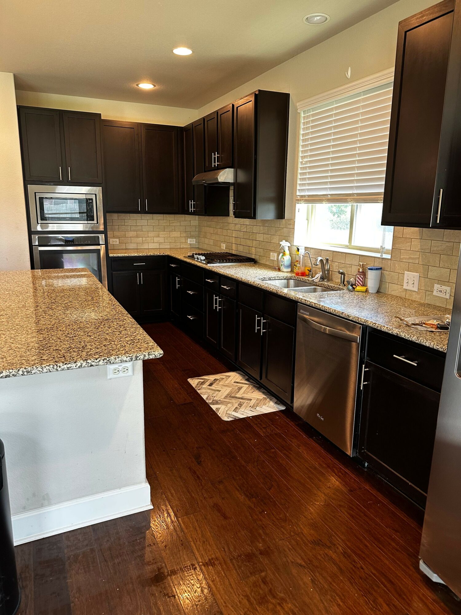 Kitchen with dark cabinets, granite countertops, stainless steel appliances, and a window with blinds, hardwood floor, and a small rug.