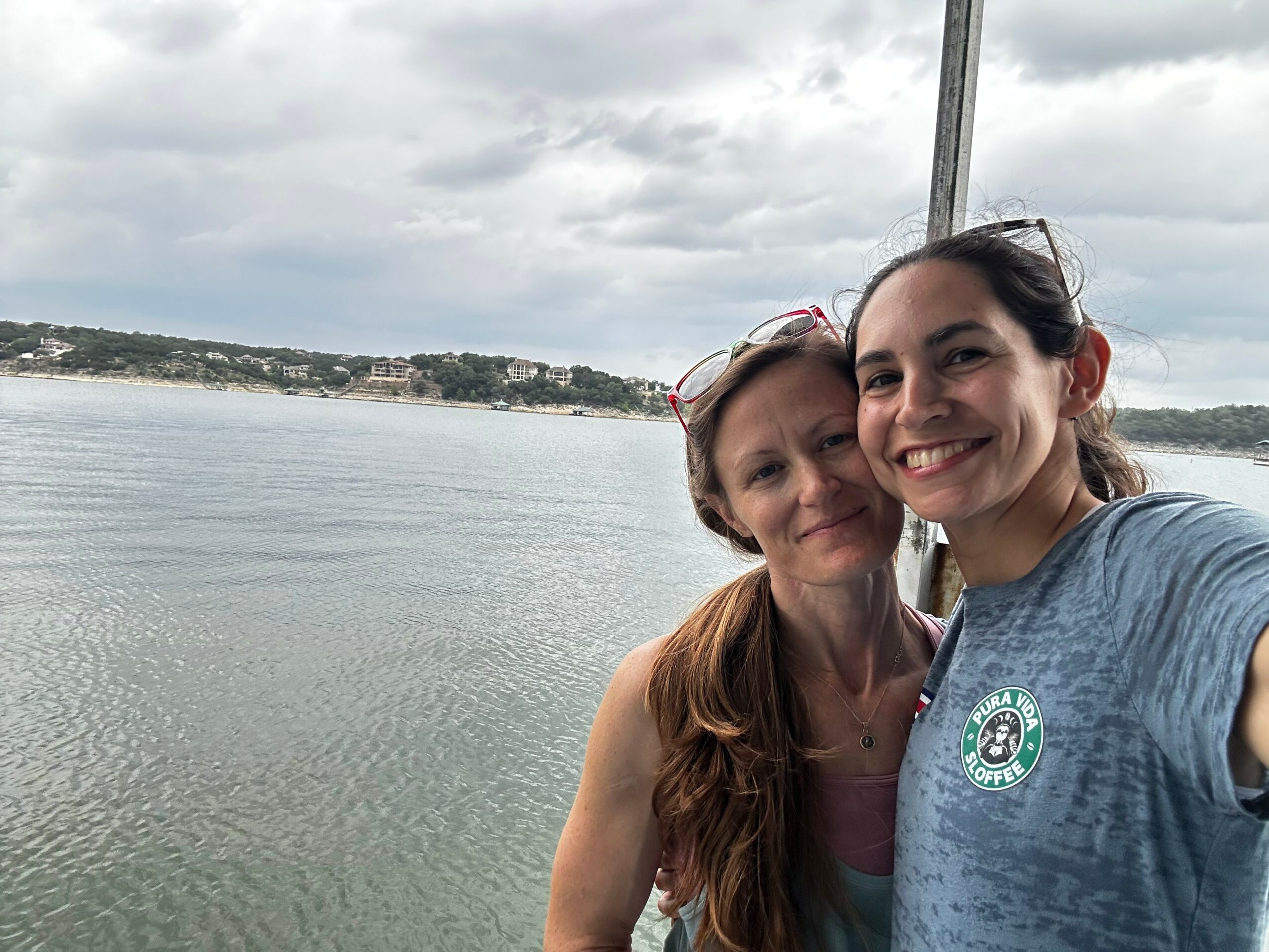 Two women smiling outdoors near water with cloudy sky, one with long red hair and glasses, the other with dark hair, wearing a gray shirt.