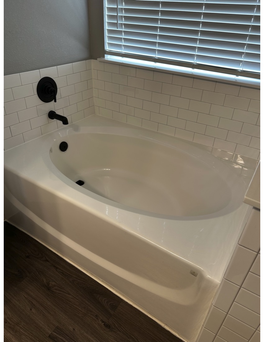 Bathtub with faucet and window with blinds above, tiled wall, and wooden floor.