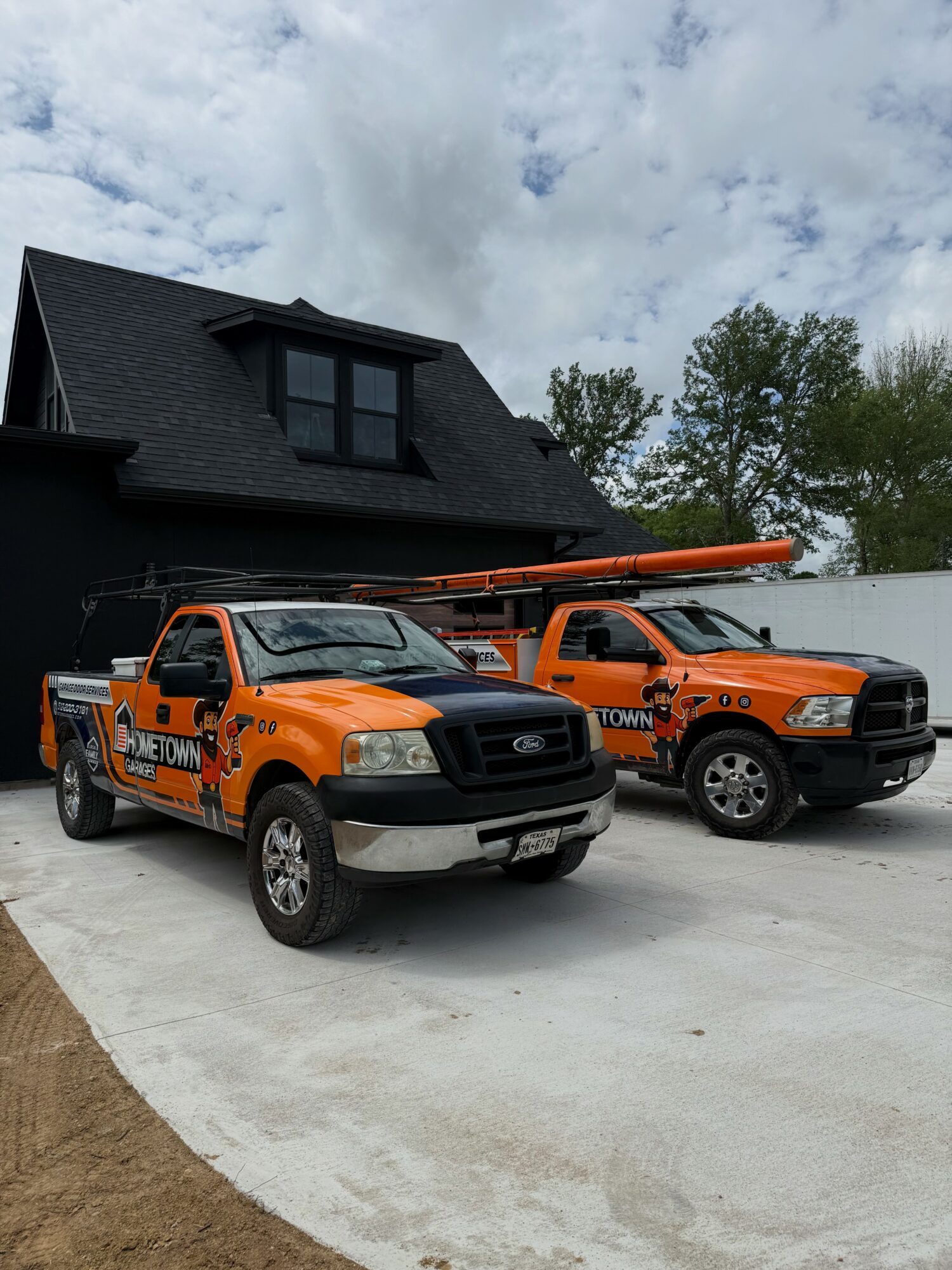 Two orange utility trucks parked in front of a black house with a sloped roof and dormer windows, under a cloudy sky.