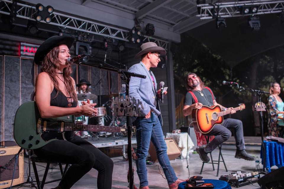 Band performing on stage with musicians playing guitar and singing, stage setup with equipment and lighting, outdoor setting.