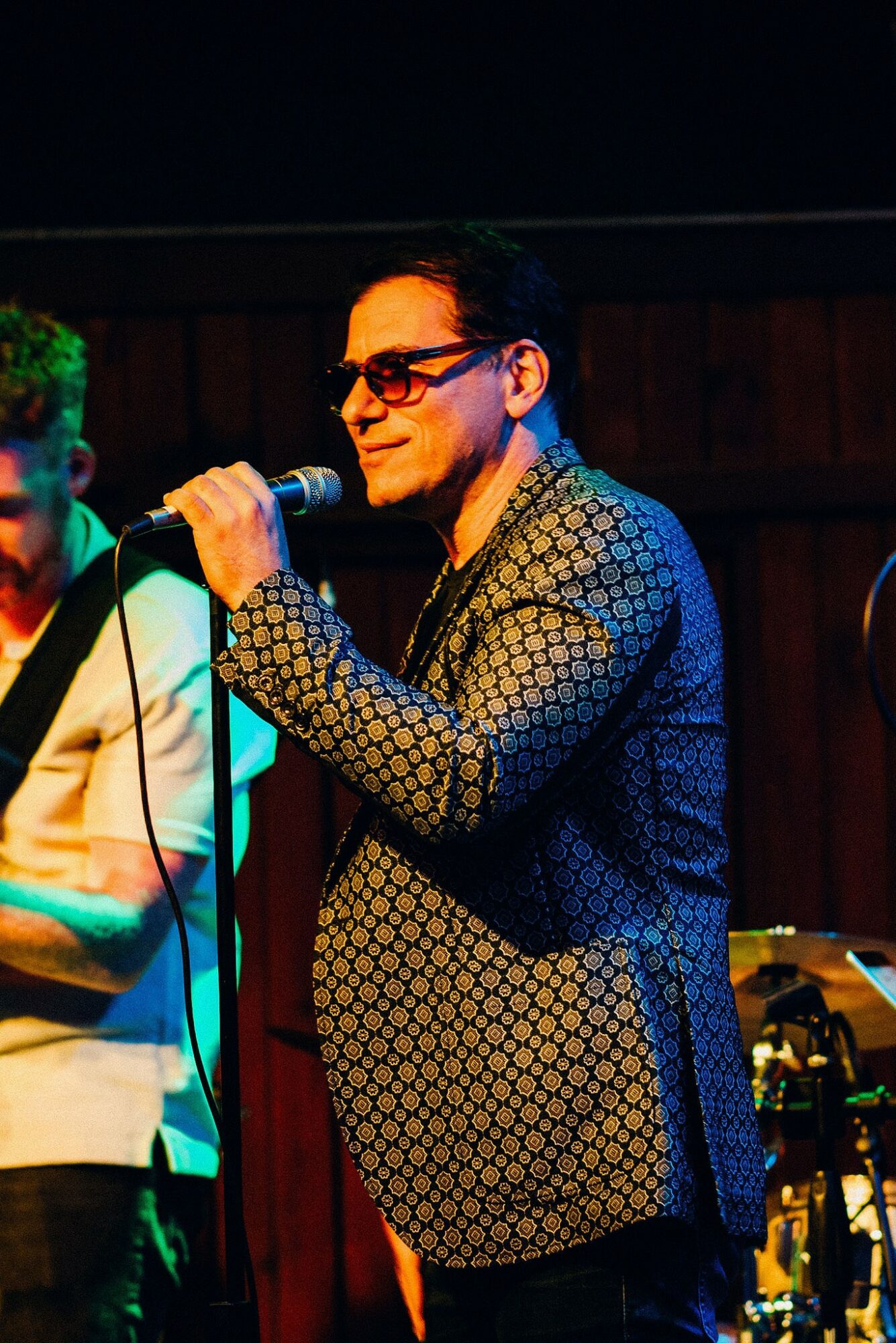Man wearing sunglasses and patterned jacket speaking into a microphone on stage.