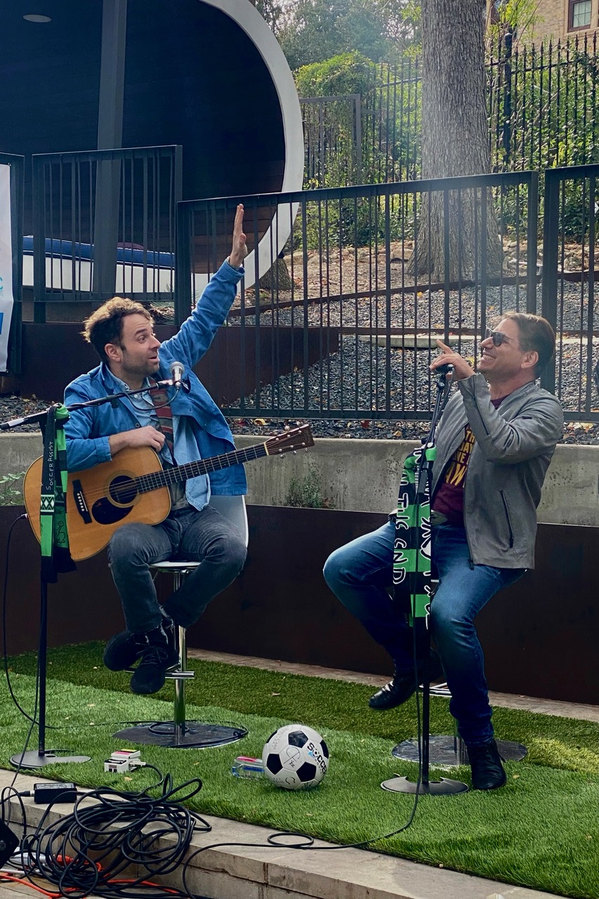 Two men perform music outdoors, one with a guitar and the other singing into a microphone, with a soccer ball nearby.