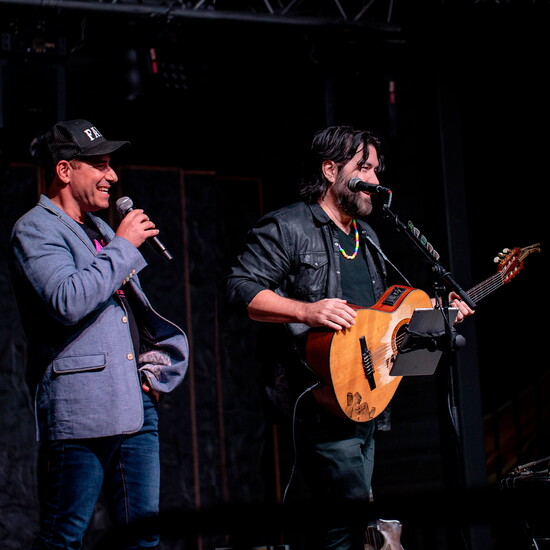 Two men on stage, one holding a microphone and the other playing a guitar, both smiling, dark background.