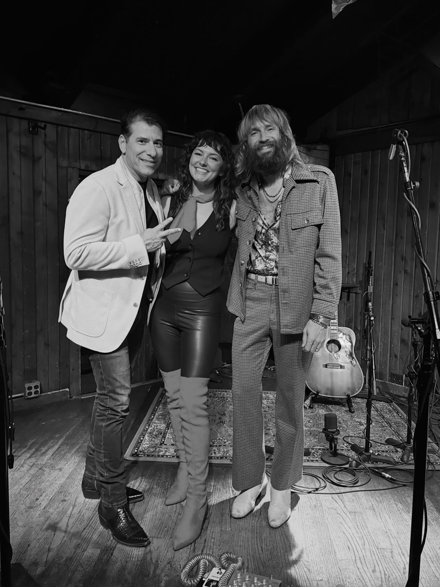 Three people standing together on a stage, smiling, with musical equipment in the background.