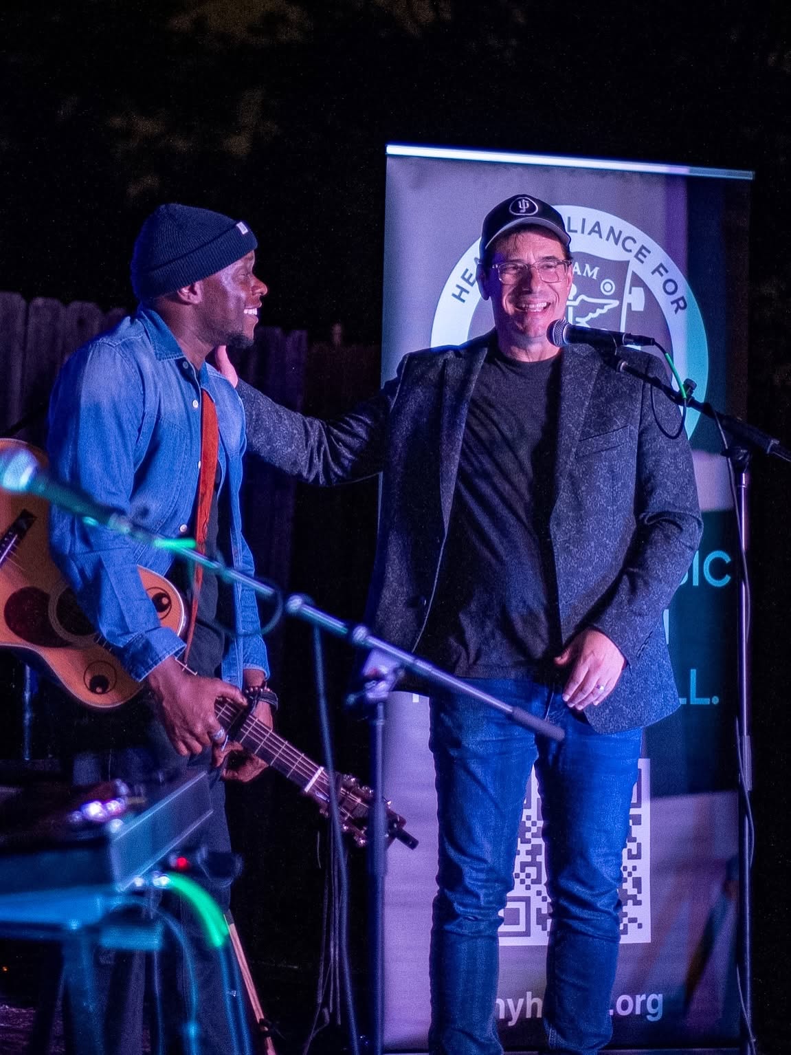 Two men on stage, one holding a guitar, smiling and talking, with a banner in the background.