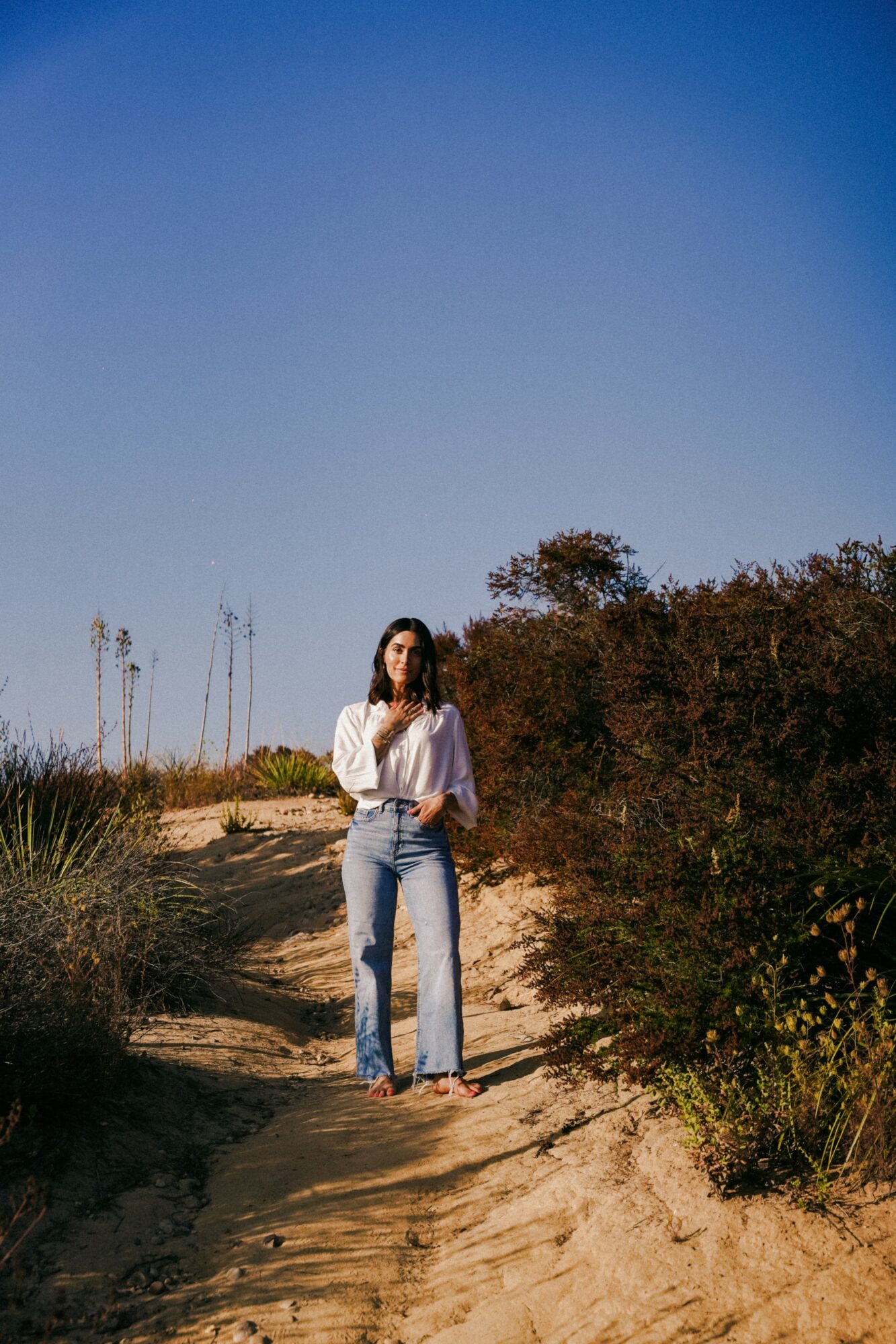Woman standing on a sandy path surrounded by bushes and plants under a clear blue sky.