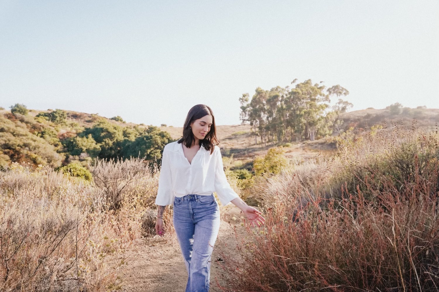 Woman walking on a dirt path in a field with dry grass and trees in the background.