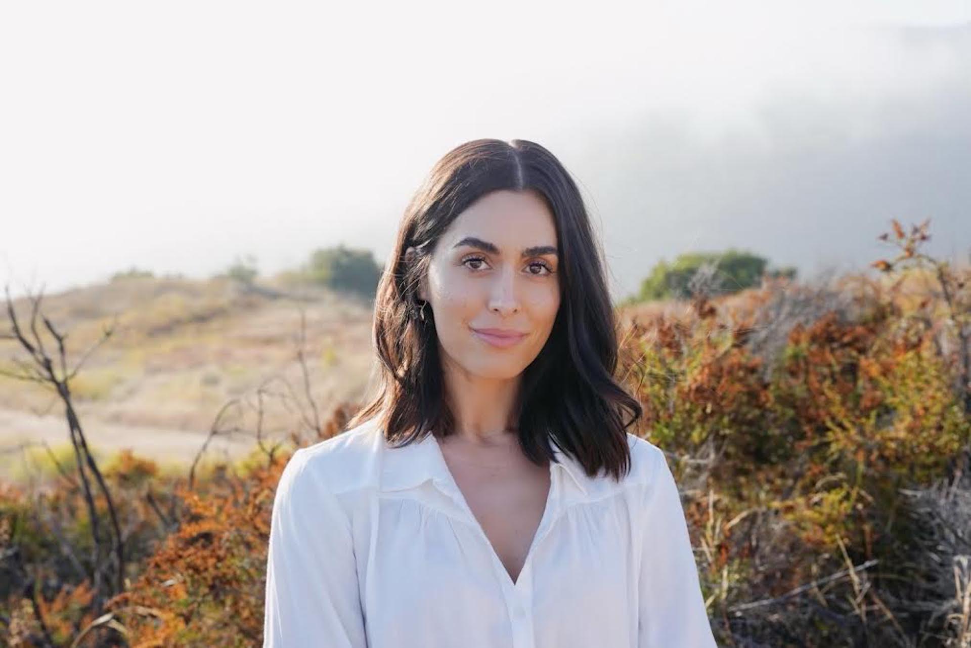 Woman with shoulder-length dark hair outdoors, wearing a white shirt, with a landscape background.