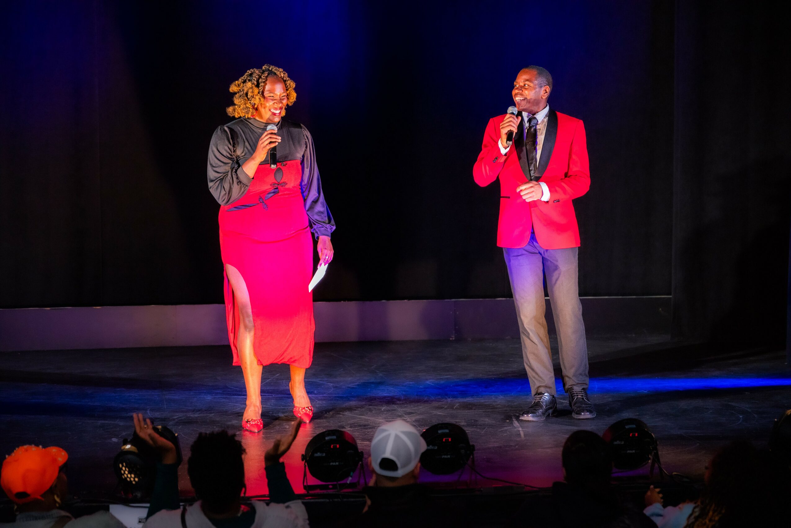 Two people stand on stage holding microphones, engaging in conversation, with an audience visible in the foreground.