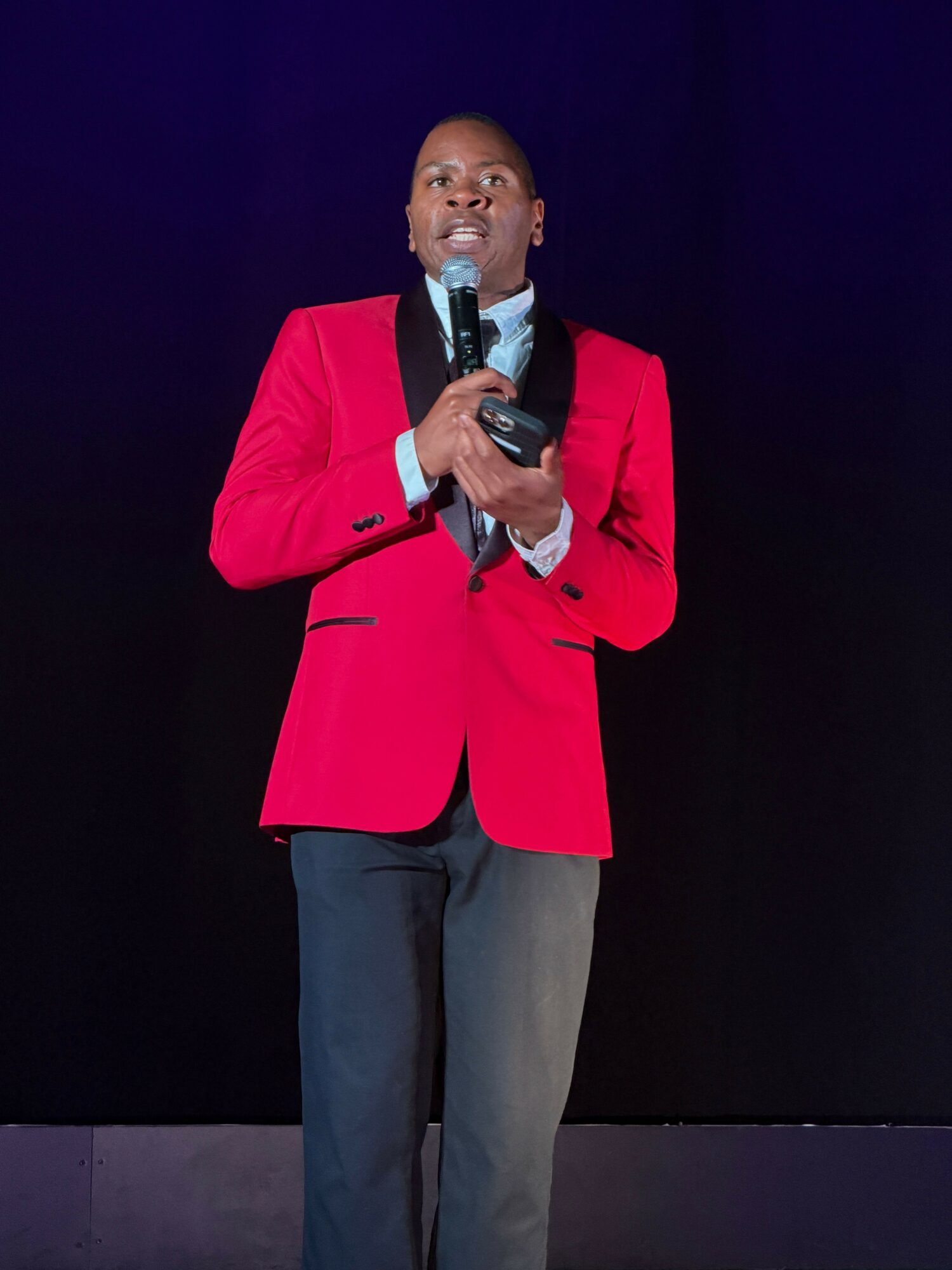 Person in red blazer holding a microphone, standing on stage with dark background.