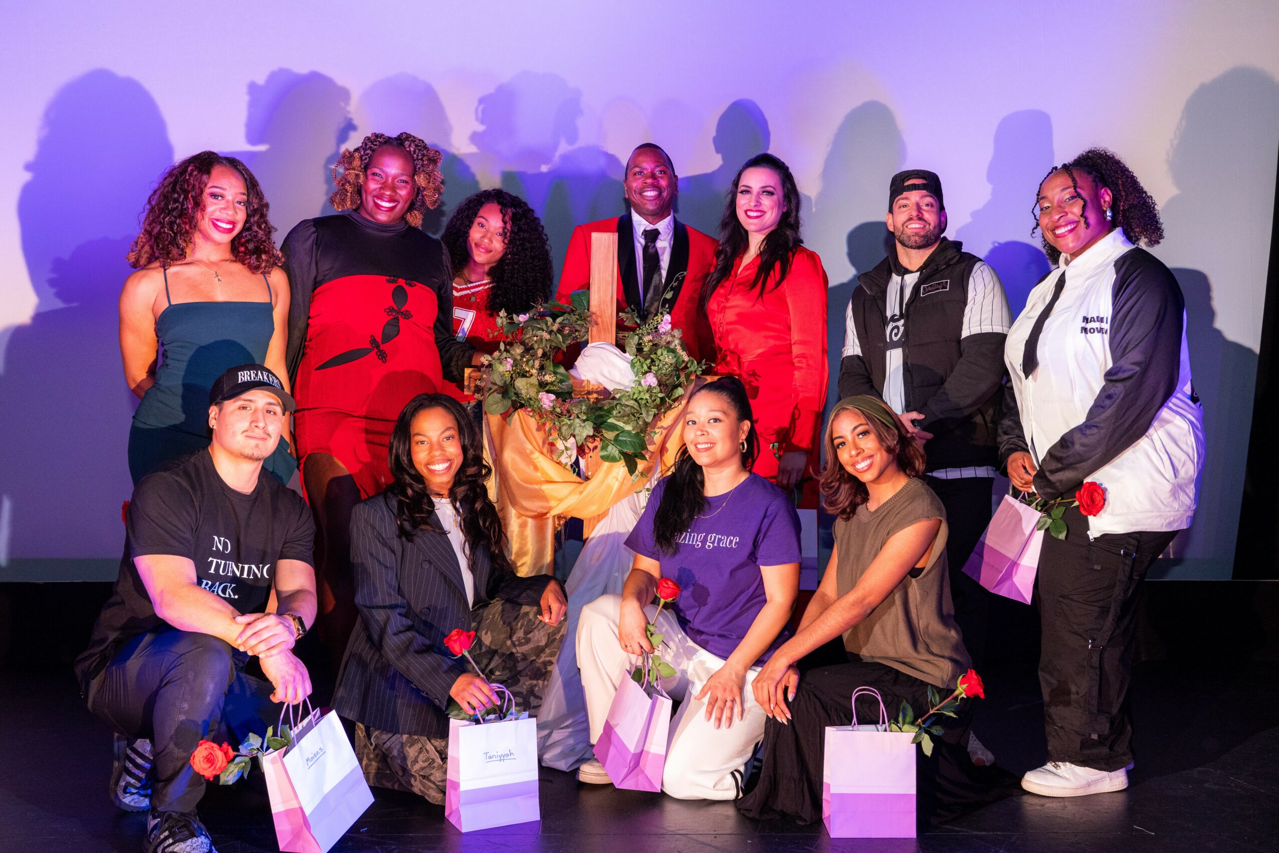 Group of ten people on stage with flowers and gift bags, smiling, in front of a plain background.