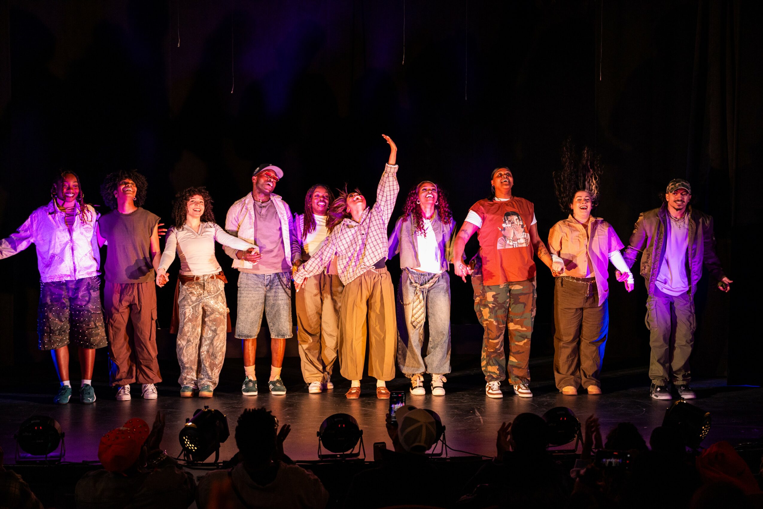 Group of people standing on stage, holding hands, with a dark background and audience silhouettes in front.