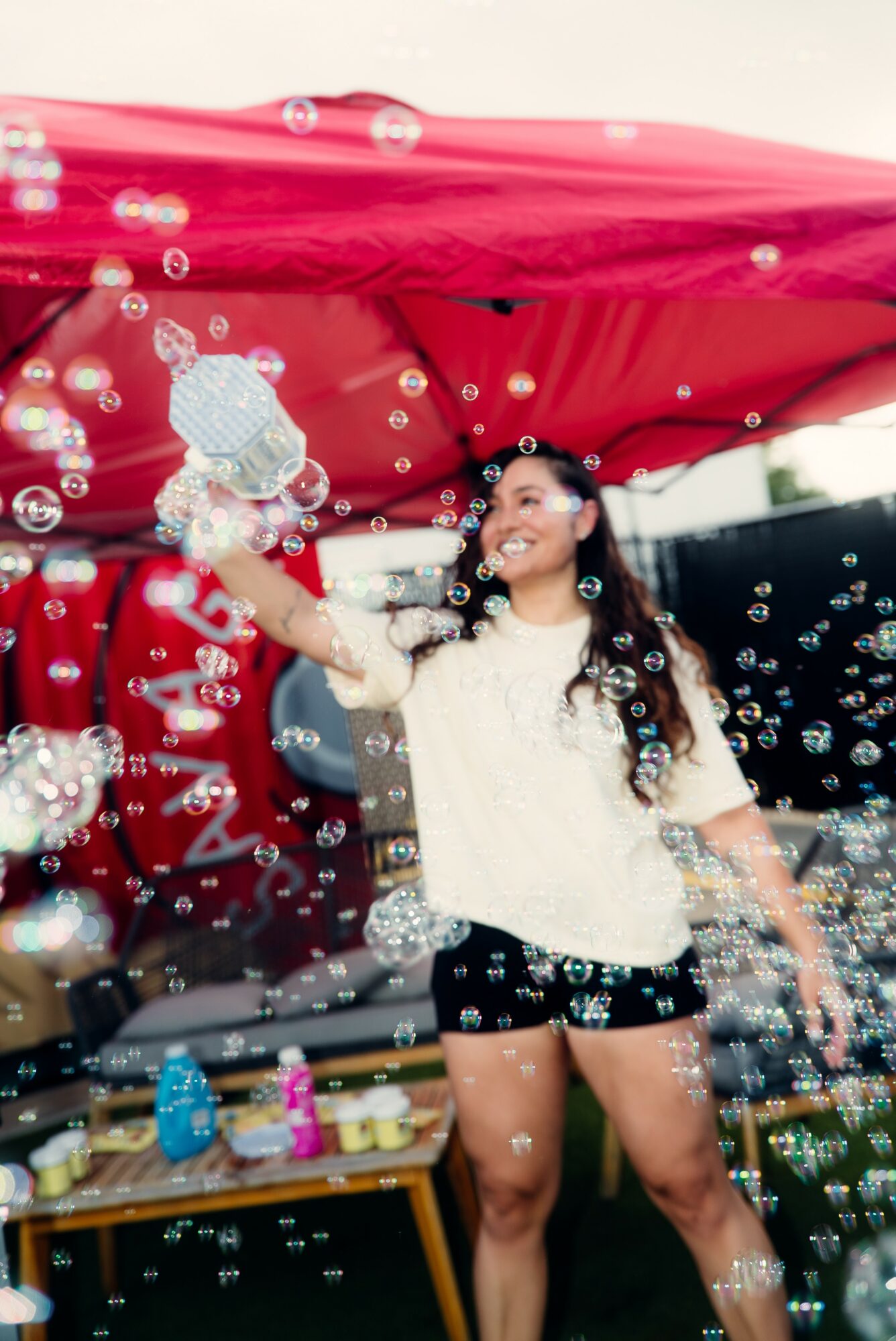 Young woman in white top and black shorts playing with bubbles outdoors, smiling, with a red canopy overhead.