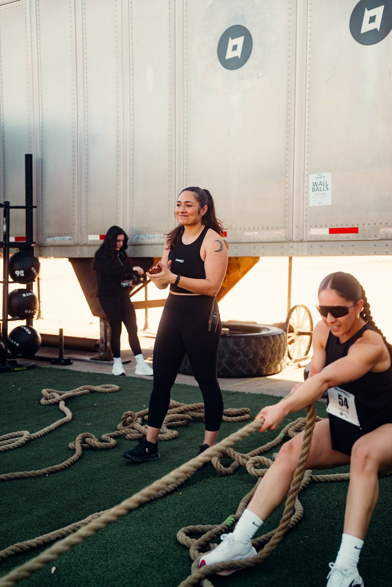 Two women in athletic clothing and sunglasses are outdoors, with one sitting and the other standing near large ropes on green ground.