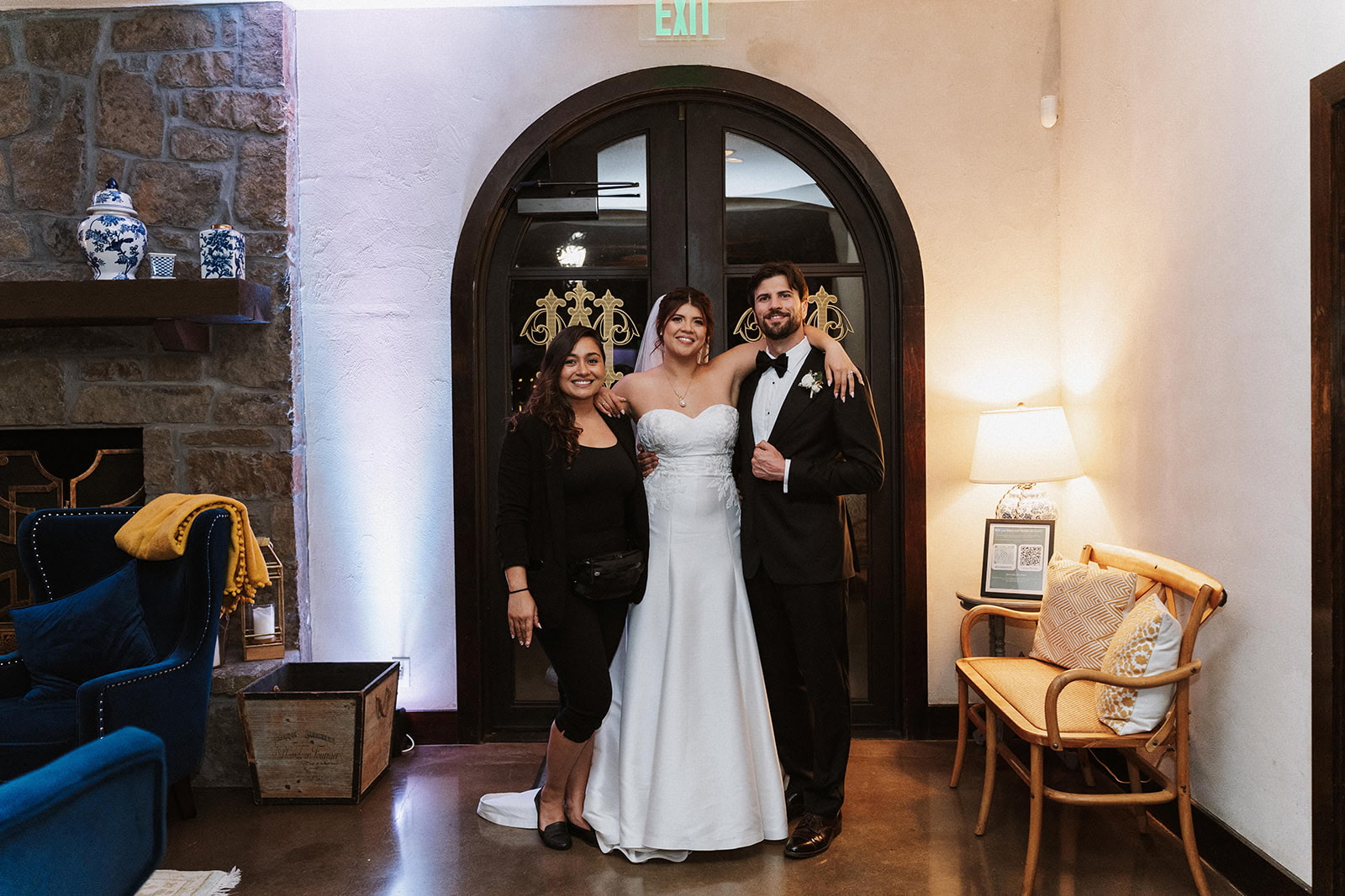 Three people, a bride in a white wedding dress and two others in formal attire, stand together inside a room with a large arched door behind them.