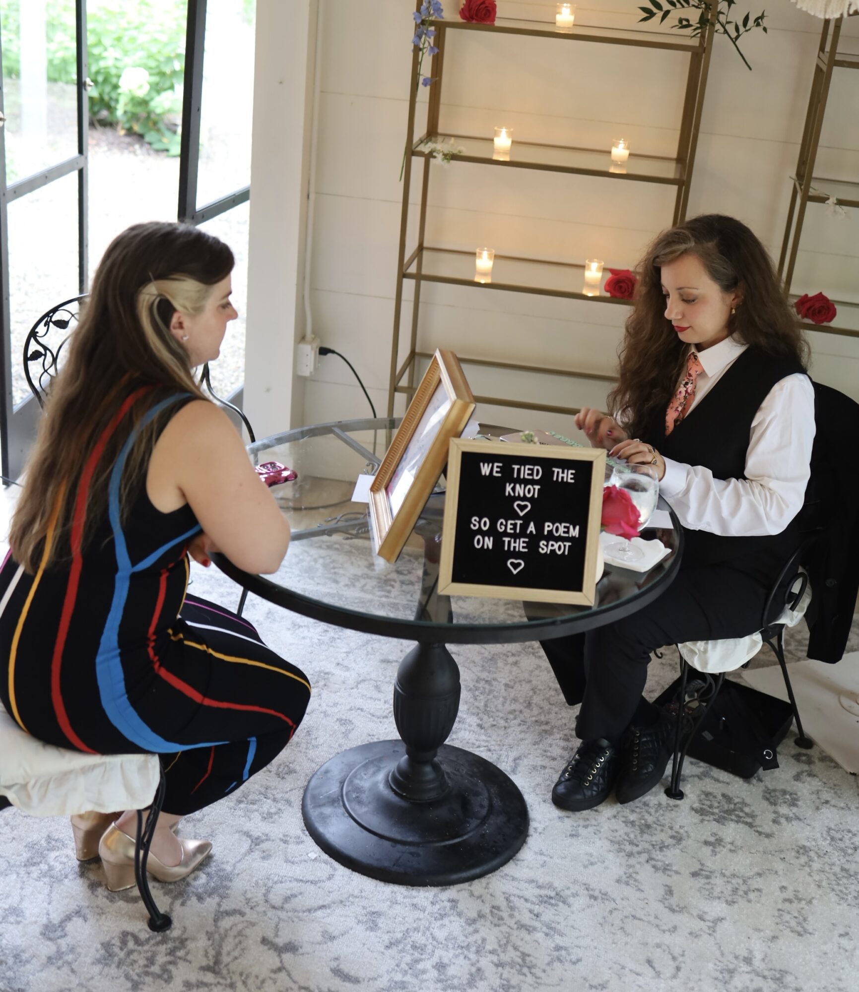 Two girls sit at a round table with a small blackboard, reading or discussing. One girl has long dark hair, the other has curly hair. Sunlight streams in through a window, and candles and flowers decorate the background.