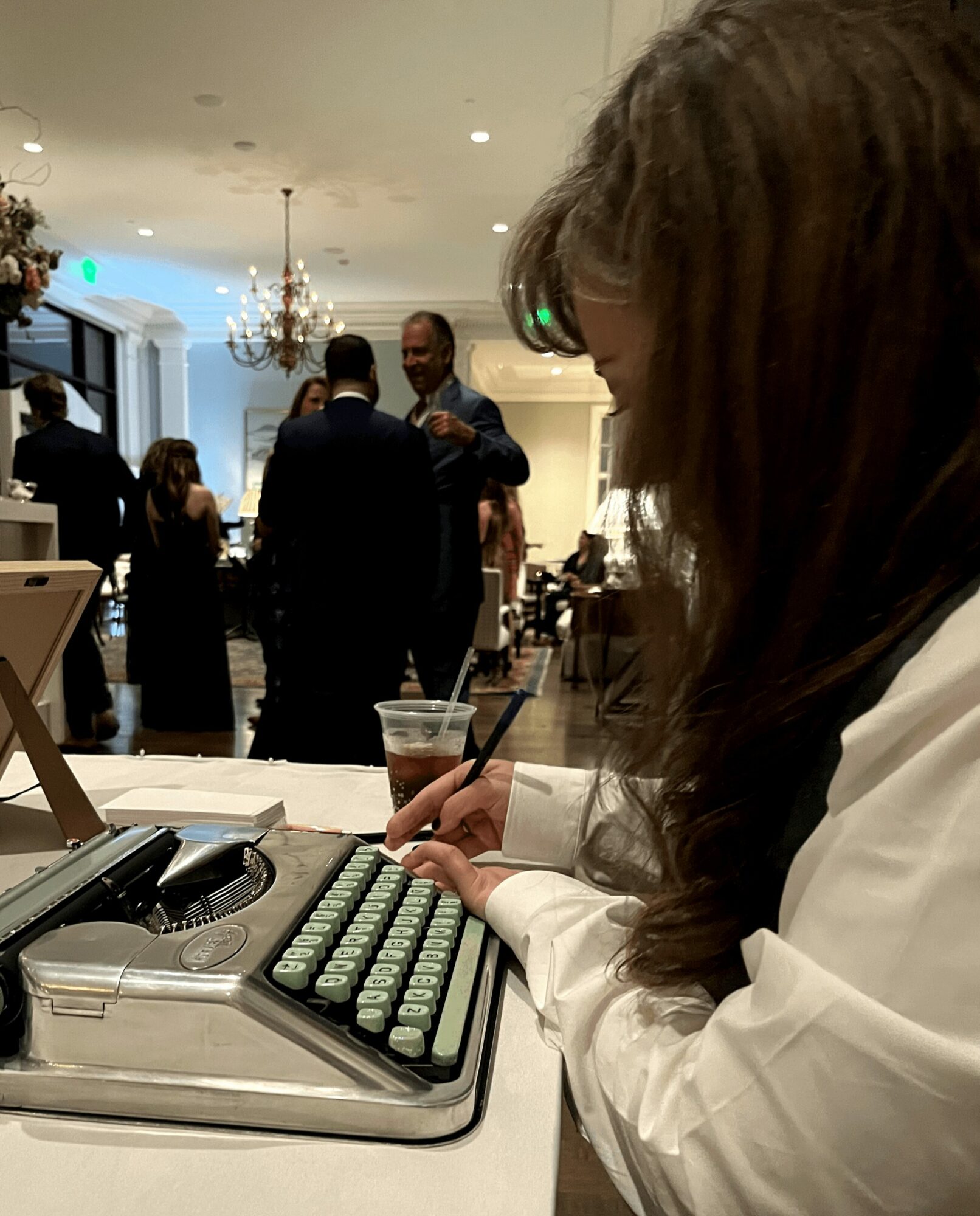 Person typing on a green keyboard at a table in a well-lit room with chandeliers and people in the background.
