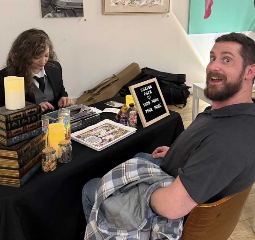Man with a beard smiling at camera seated at table with books, snacks, and a sign, woman sitting behind table, art on wall.
