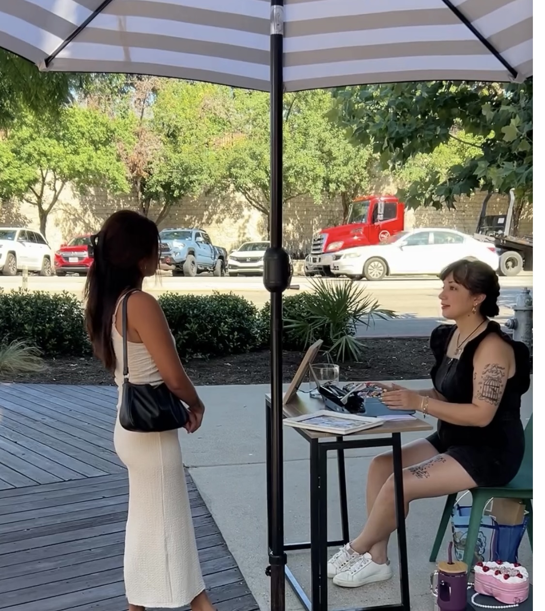 Two women converse outdoors under an umbrella, one seated at a table with a laptop, the other standing nearby, with trees and cars in background.