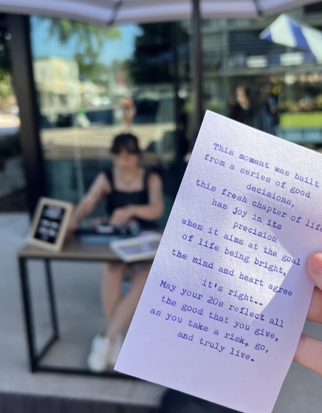A handwritten note held in front of a blurred outdoor cafe scene with a woman sitting at a table.