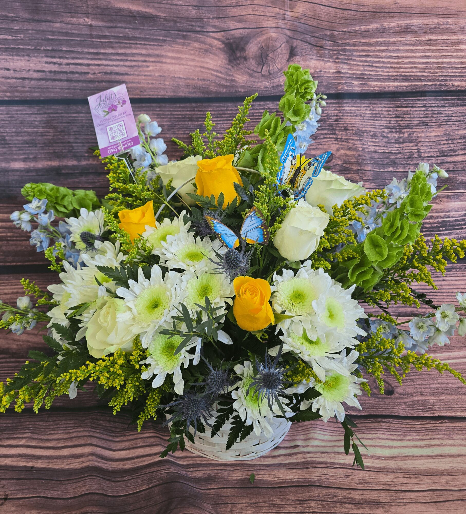 Flower arrangement with yellow roses, white daisies, and greenery in a glass vase on wooden surface.
