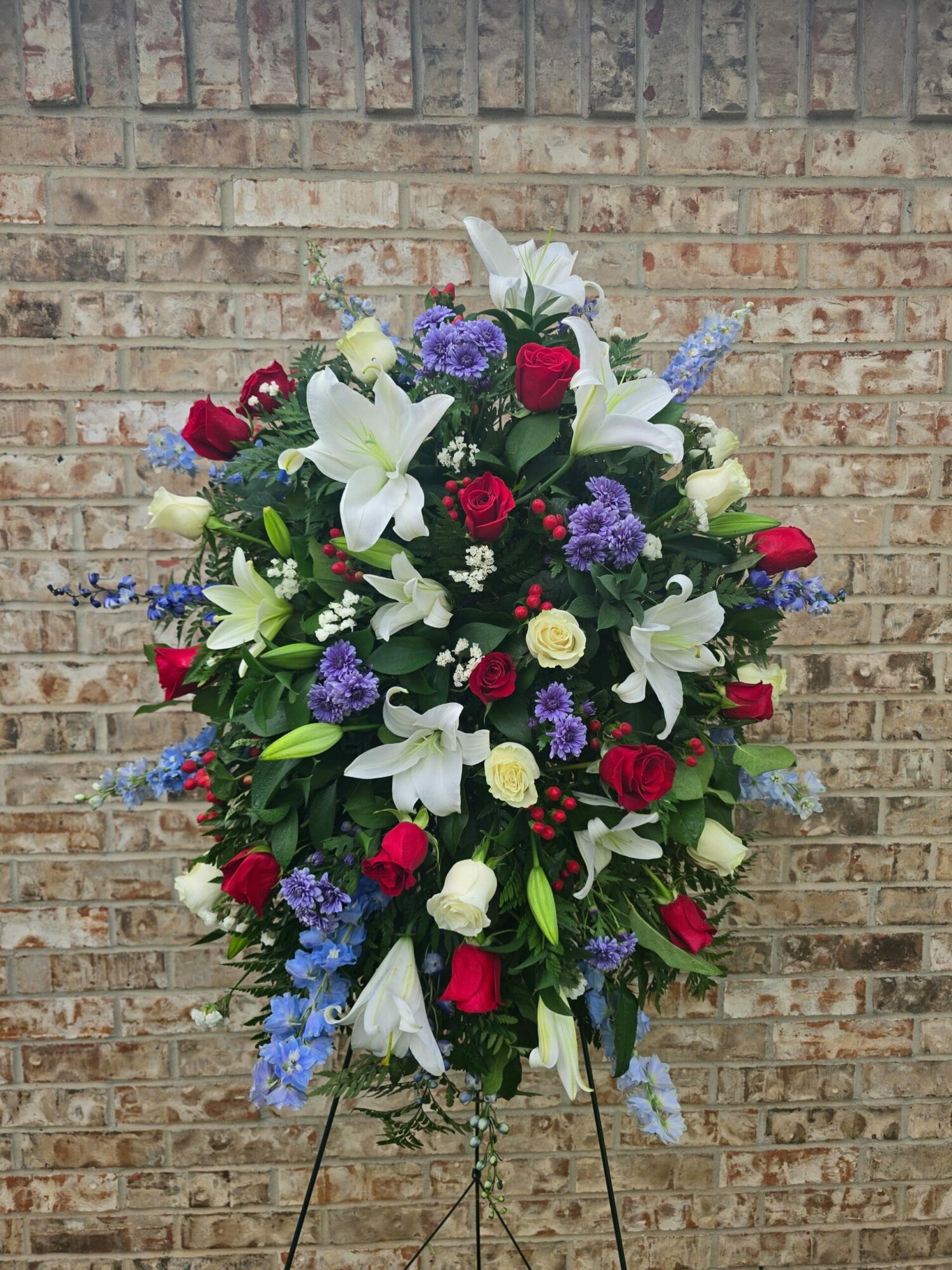 A large floral arrangement with white lilies, red roses, purple and white flowers, and green foliage, against a brick wall.