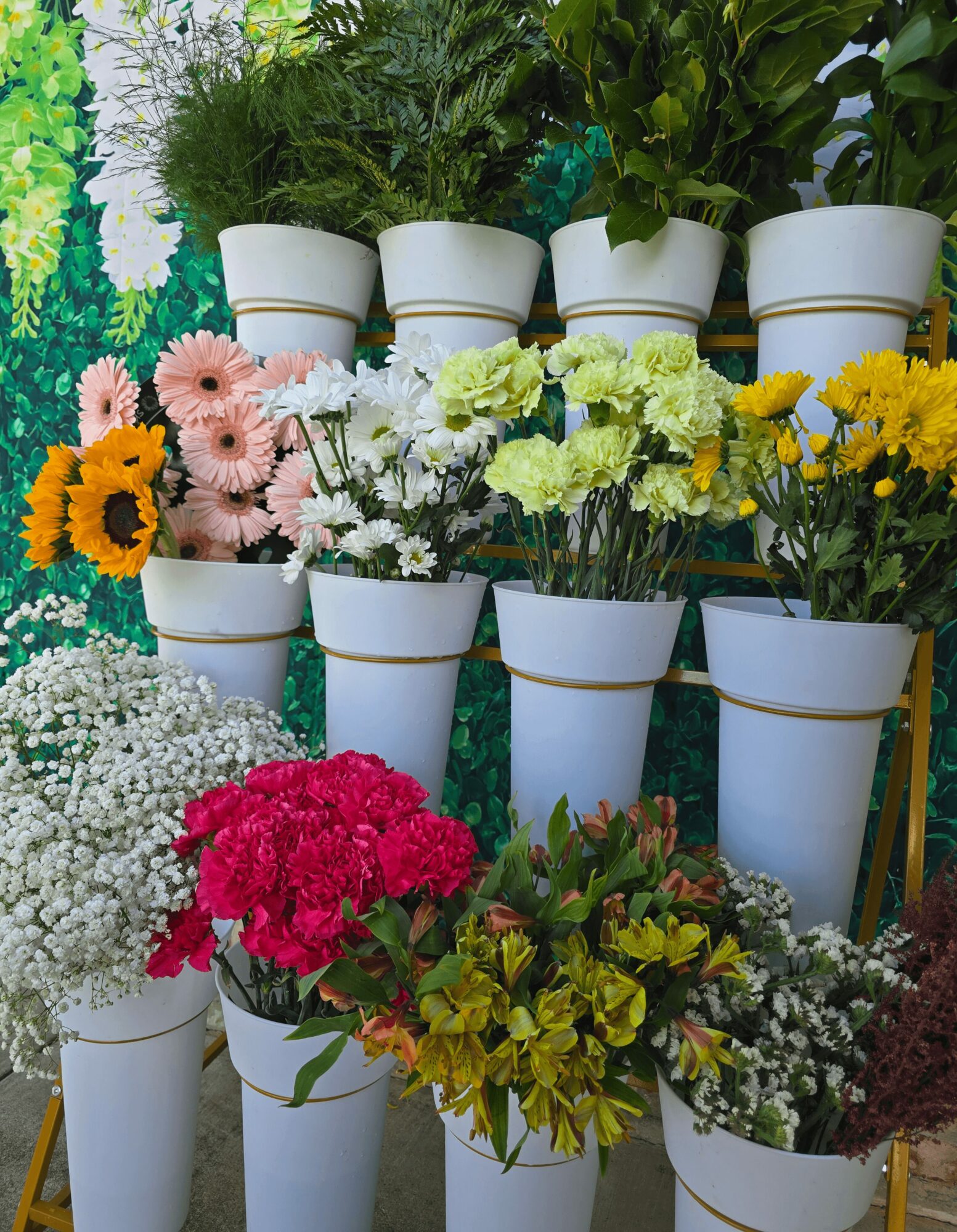 Multiple potted flowers arranged on a stand with a green leafy background, including sunflowers, daisies, and other colorful blooms.