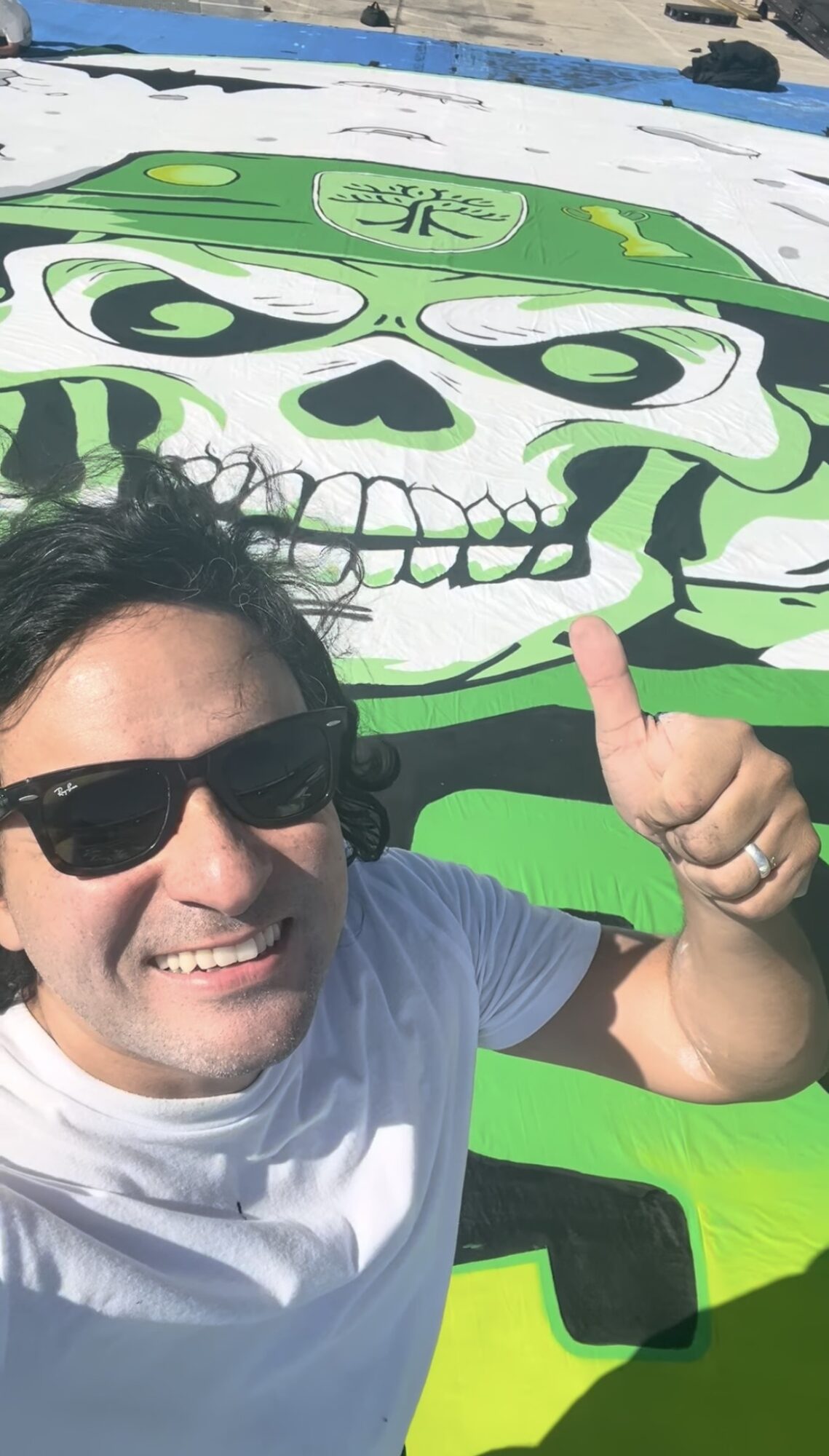 Person with sunglasses smiling and giving a thumbs-up in front of a colorful skull mural outdoors.