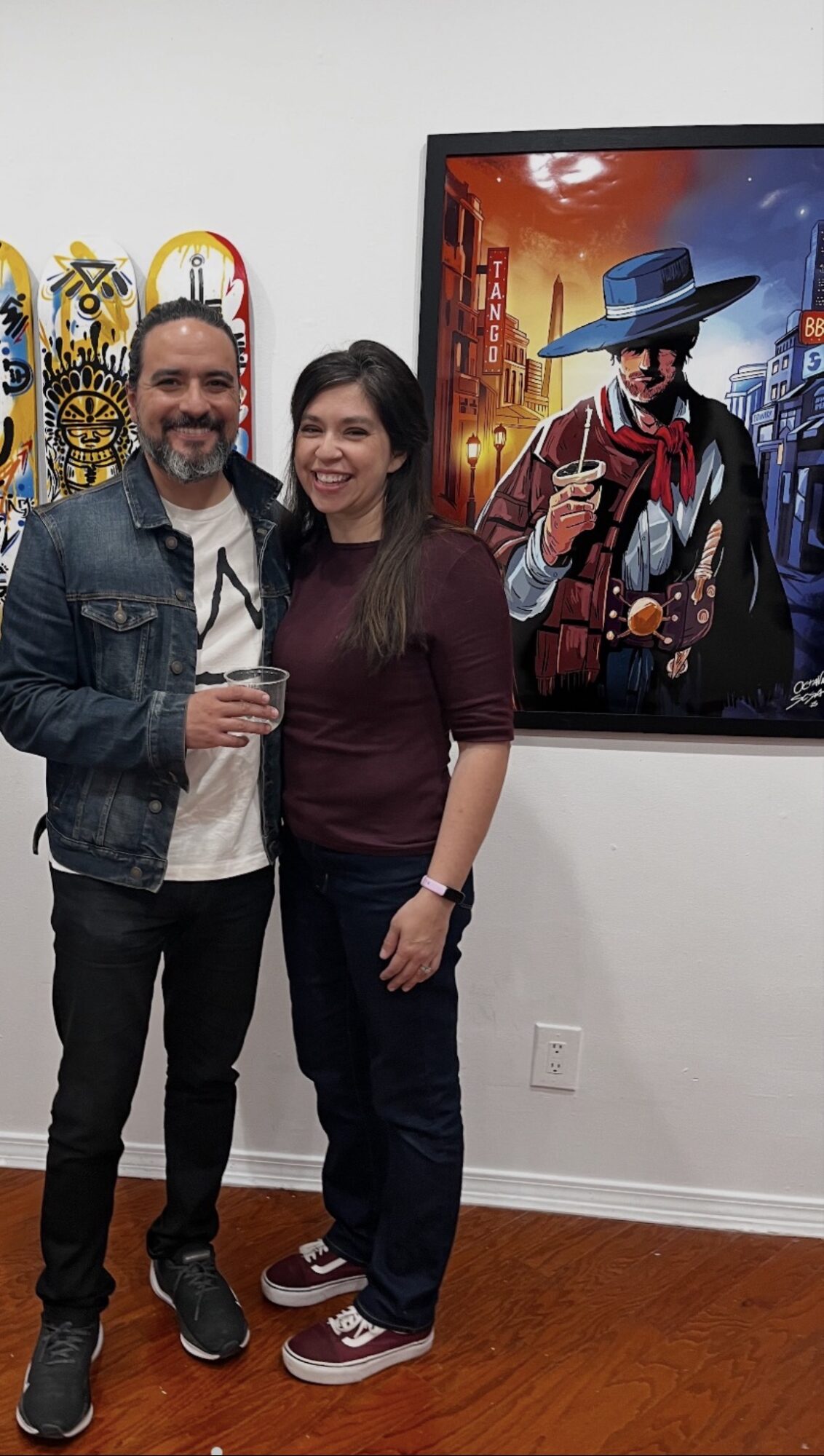 Two people standing indoors, smiling, with a colorful poster of a character in a hat and coat on the wall behind them.