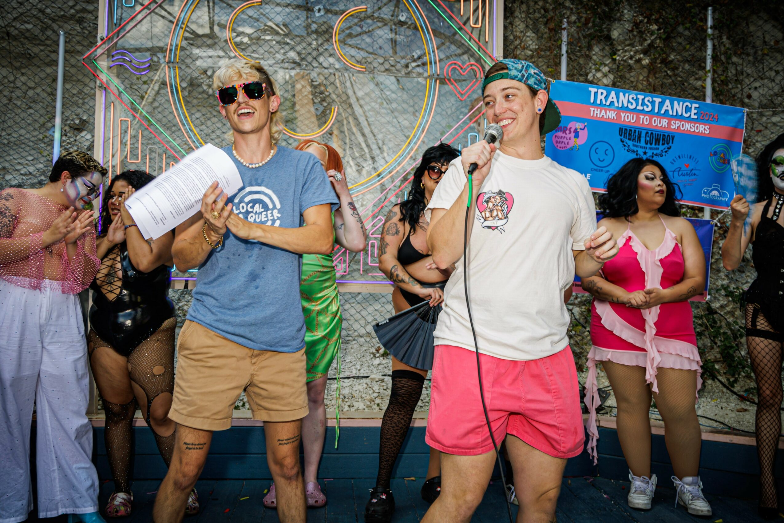Group of people on stage, some holding microphones, with a colorful backdrop and a blue banner reading 'TRANSISTANCE'.