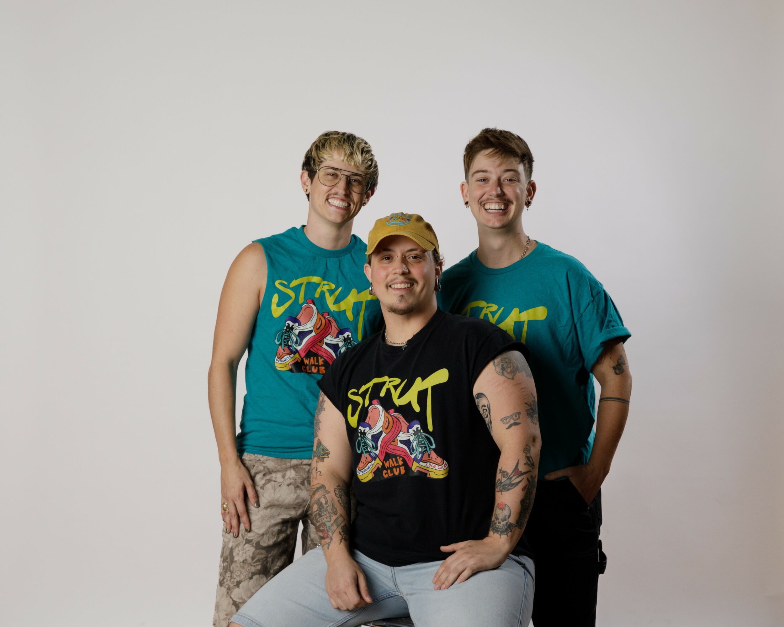 Three smiling people standing together against a plain background, two wearing teal shirts and one in a black shirt, all with casual clothing.