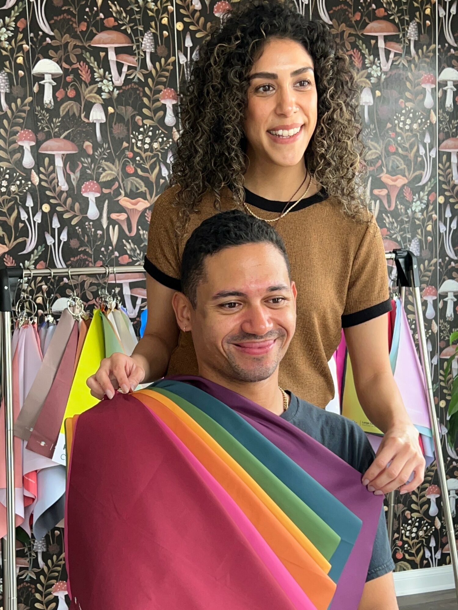 A woman with curly hair and a man with short hair smiling, holding colorful fabric, in a shop with patterned wallpaper.