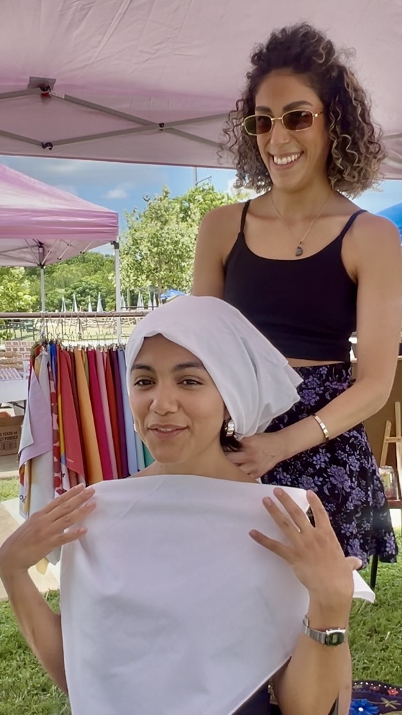 Two women outdoors, one with curly hair and sunglasses, the other with a headscarf, smiling near colorful clothing displays.