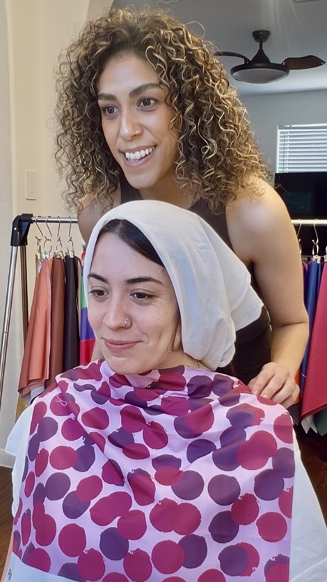Two women, one with curly hair, smiling, one with a headscarf, in a clothing store, colorful clothes in background.