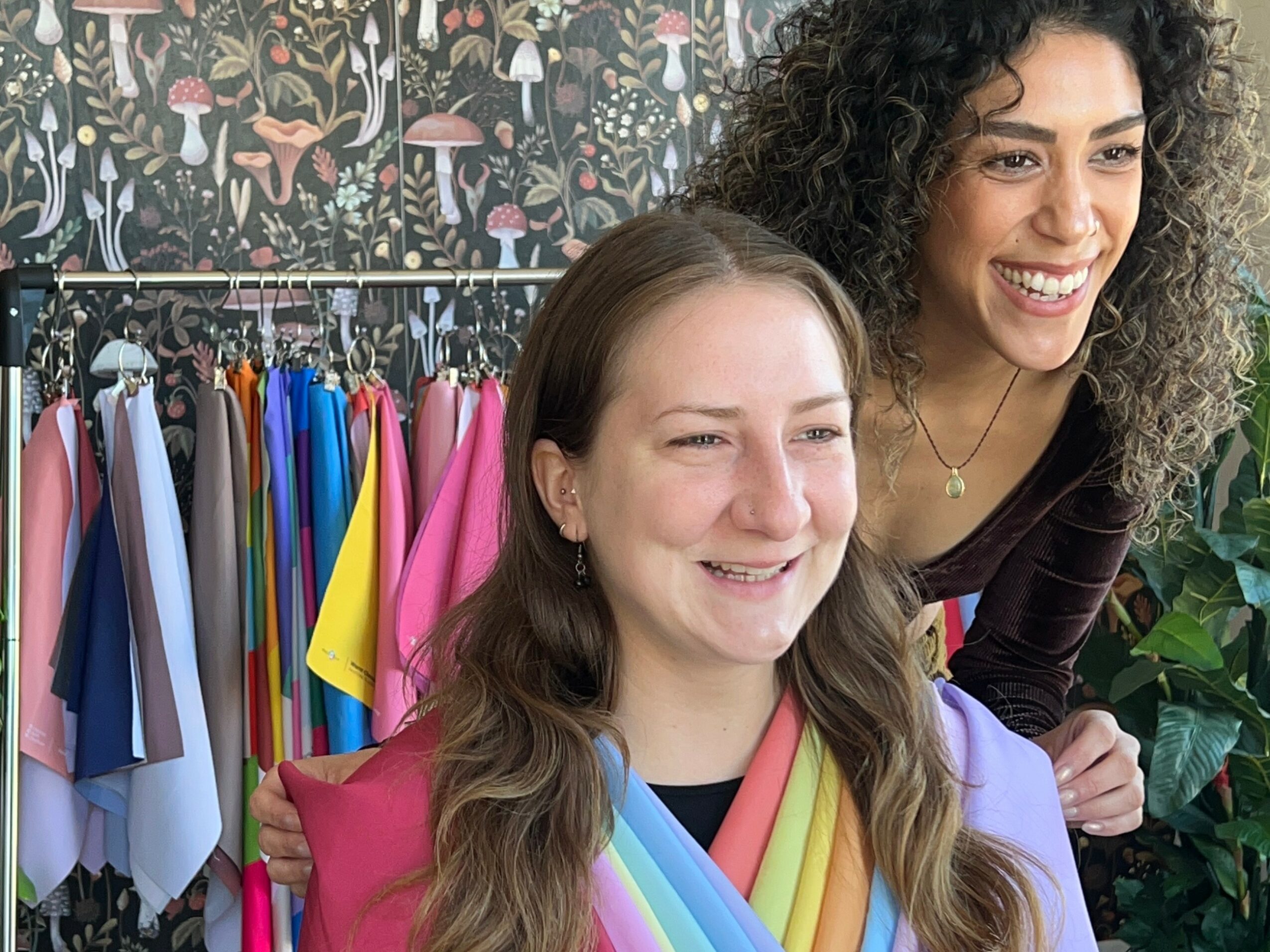Two women smiling, one with long brown hair, the other with curly hair, in a colorful clothing store.