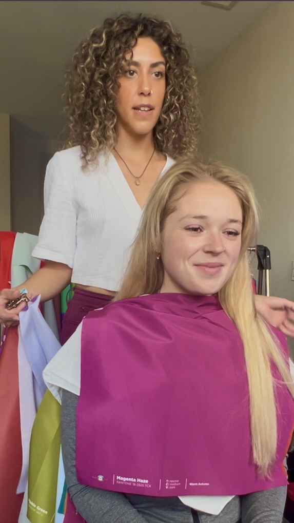 Woman with curly hair standing behind a seated woman with long blonde hair, wearing a pink cape, in a room.