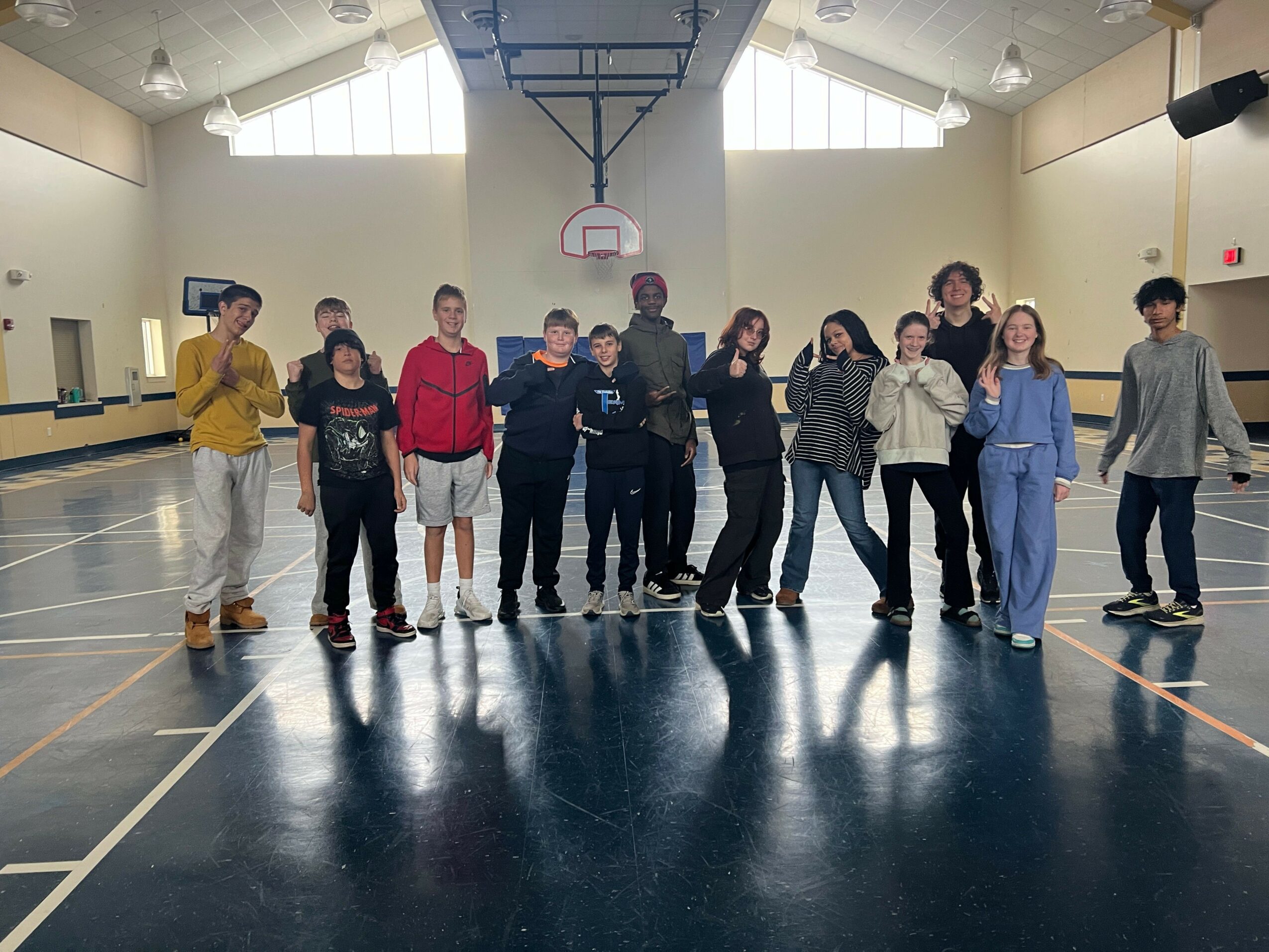 Group of people standing in a gymnasium, facing the camera, with a basketball hoop behind them, and reflections on the shiny floor.