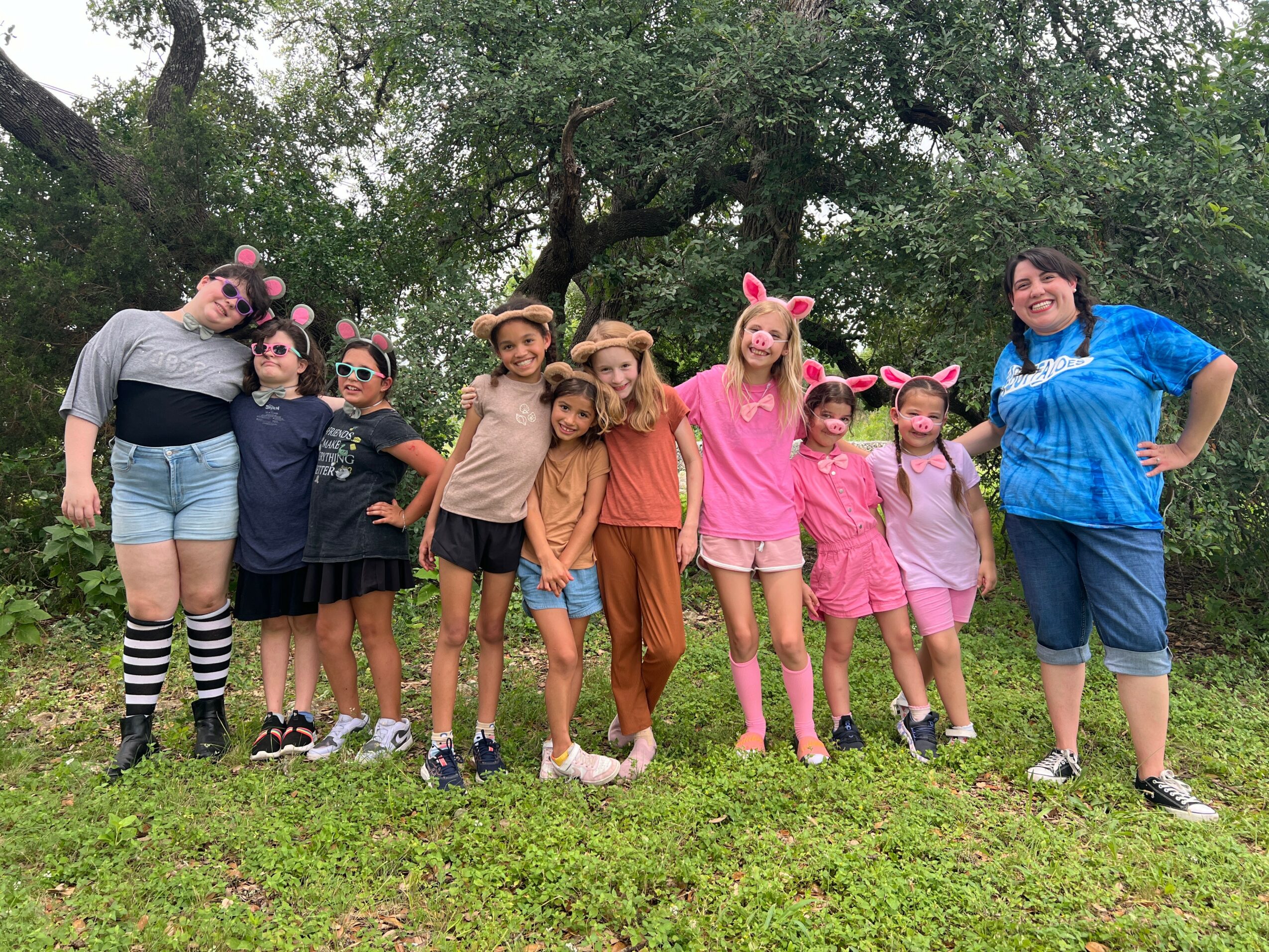 Group of children and an adult outdoors, some wearing animal ears, standing on grass with trees in background.
