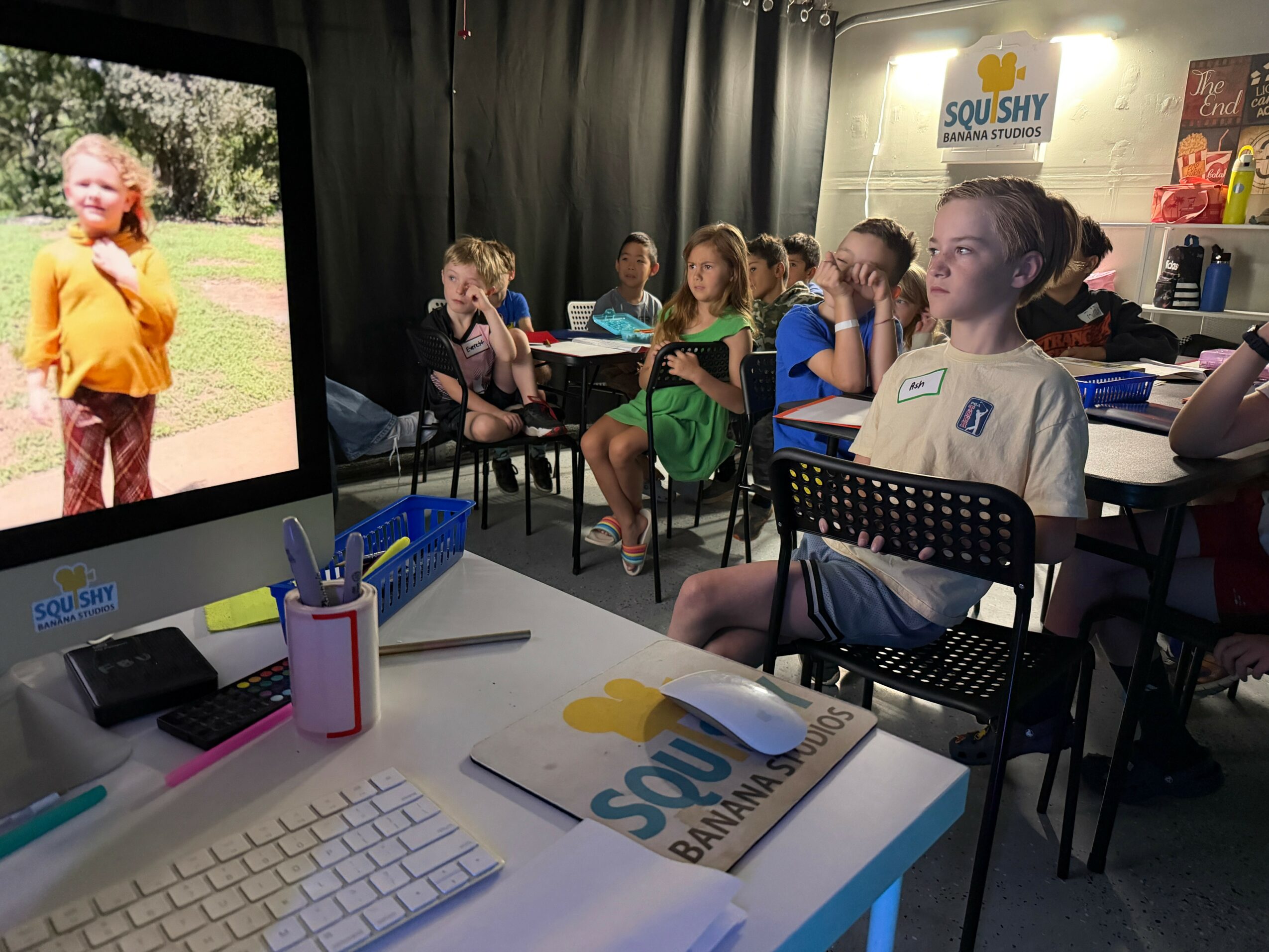 Children seated in a classroom watching a girl on a screen, with educational materials on the desk.