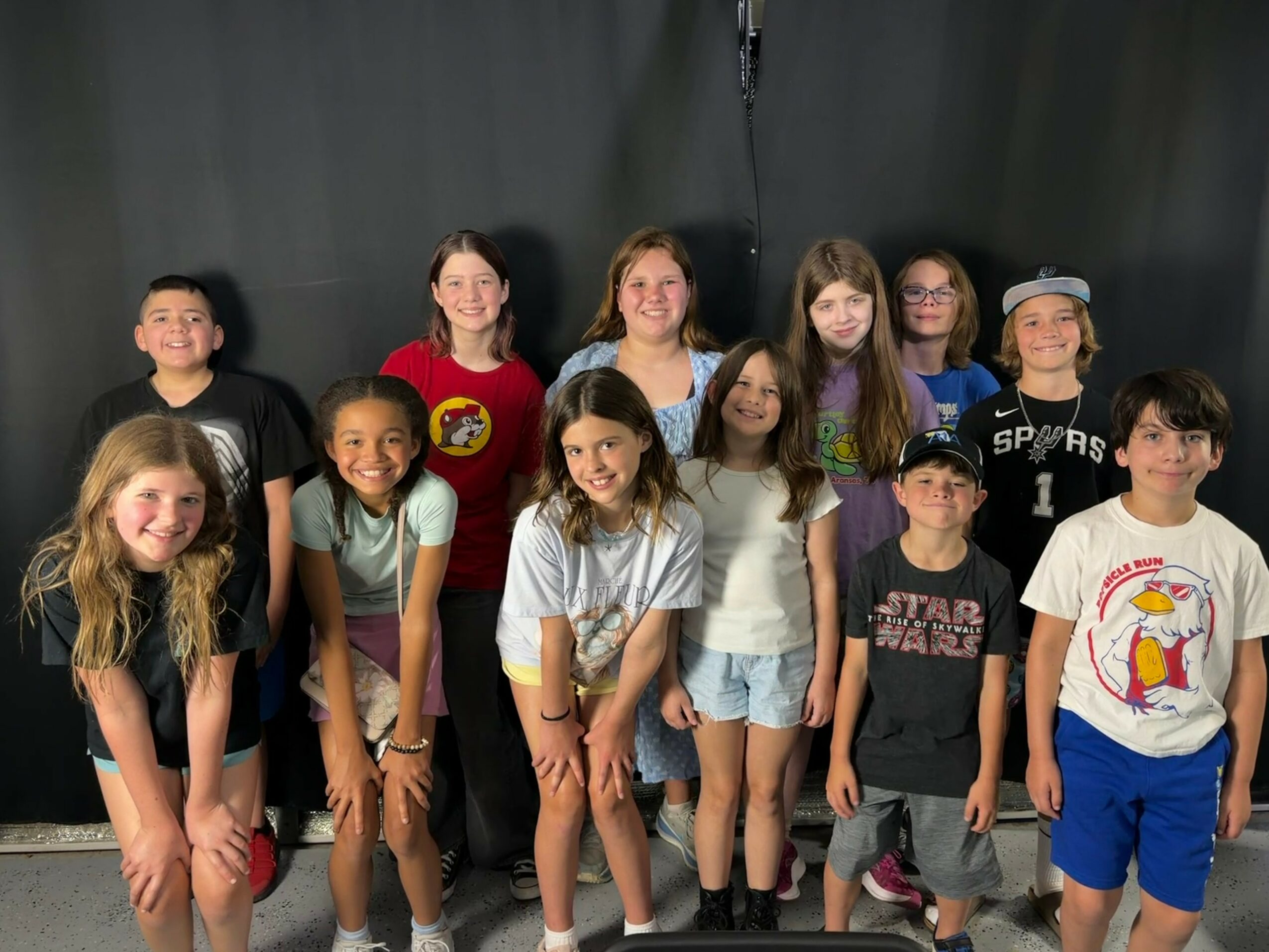 Group of twelve children standing in front of a black background, smiling, wearing casual clothes.