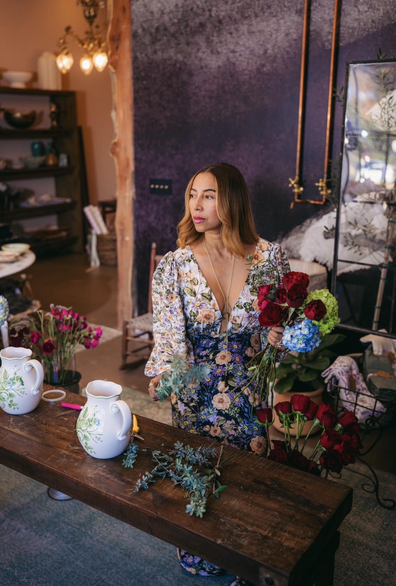 Woman with long hair in floral dress standing near a table with flowers and teapots, interior setting with purple wall and shelves.