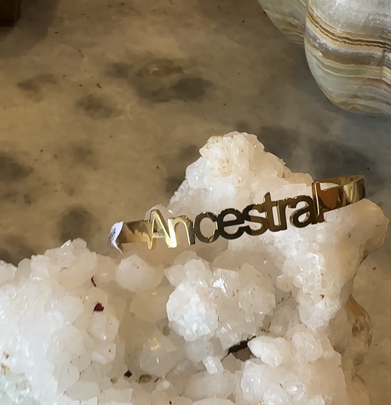 Gold-colored bracelet with the word 'Ancestral' rests on a cluster of white salt crystals, with a blurred background.