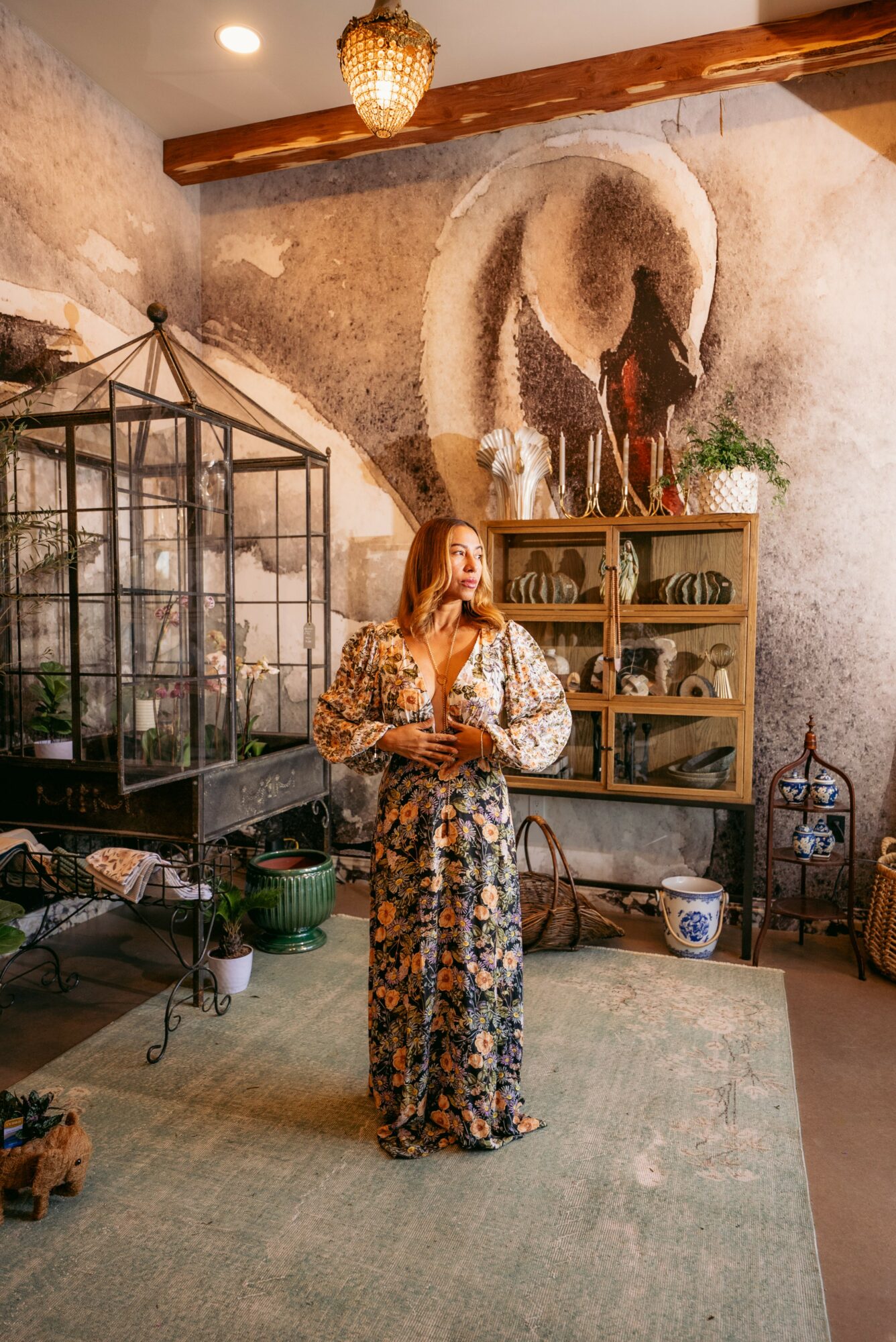 Woman in patterned dress and face mask stands in rustic room with wall art, shelves, and plants, holding a drink.