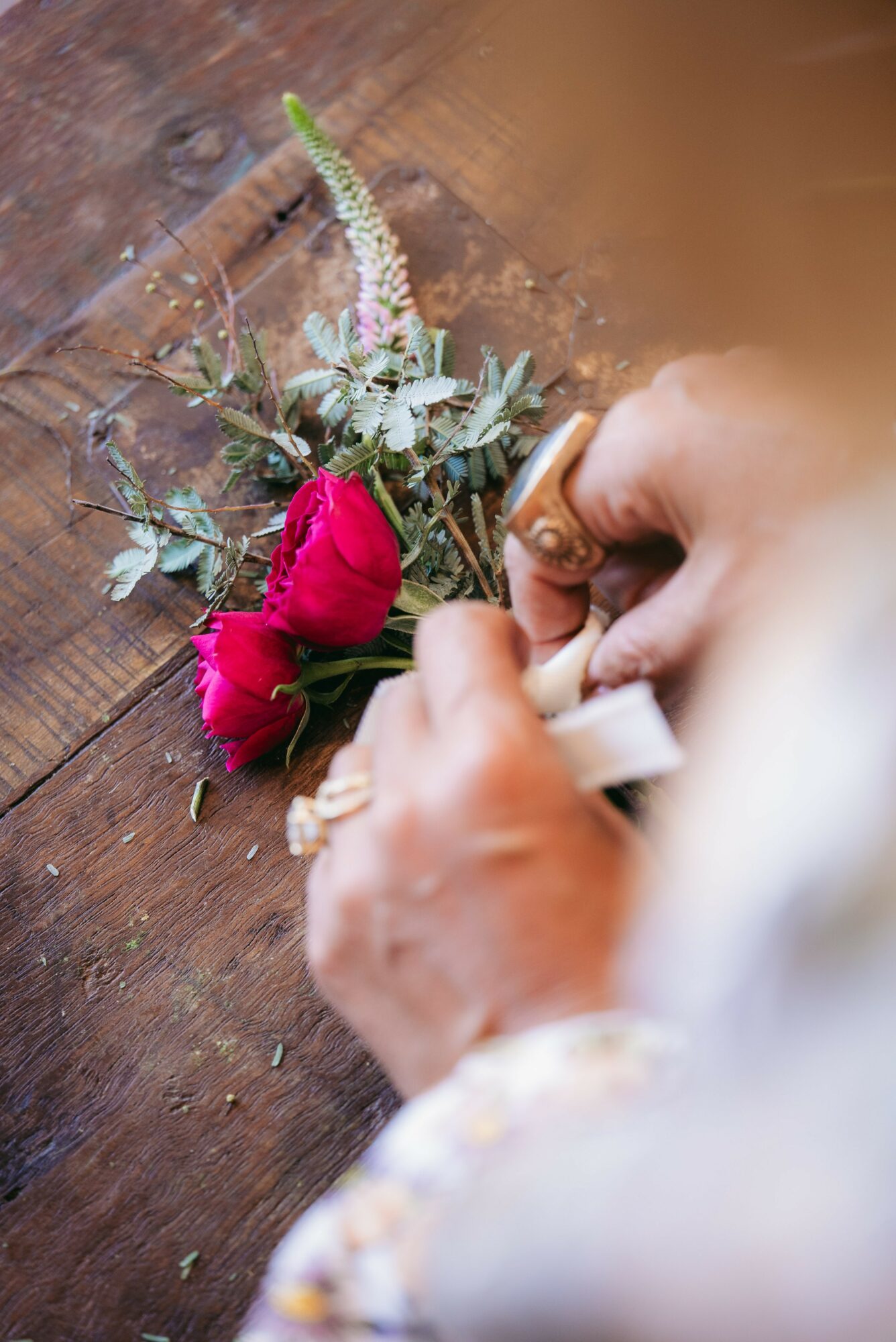 Person's hands arranging a small bouquet of pink flowers and greenery on a wooden surface.