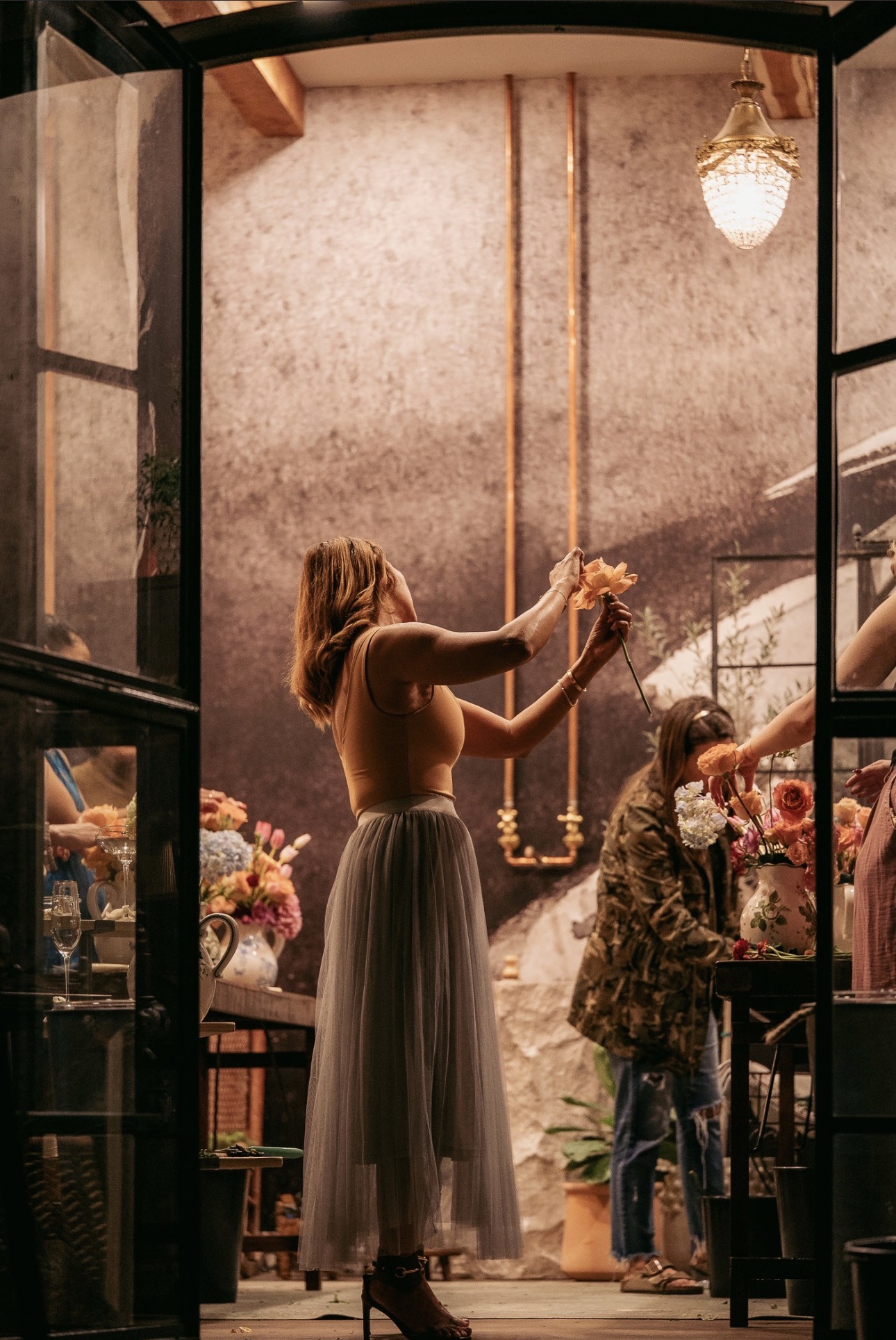 Woman in a long skirt reaching for a hanging object inside a room with flowers and a chandelier.