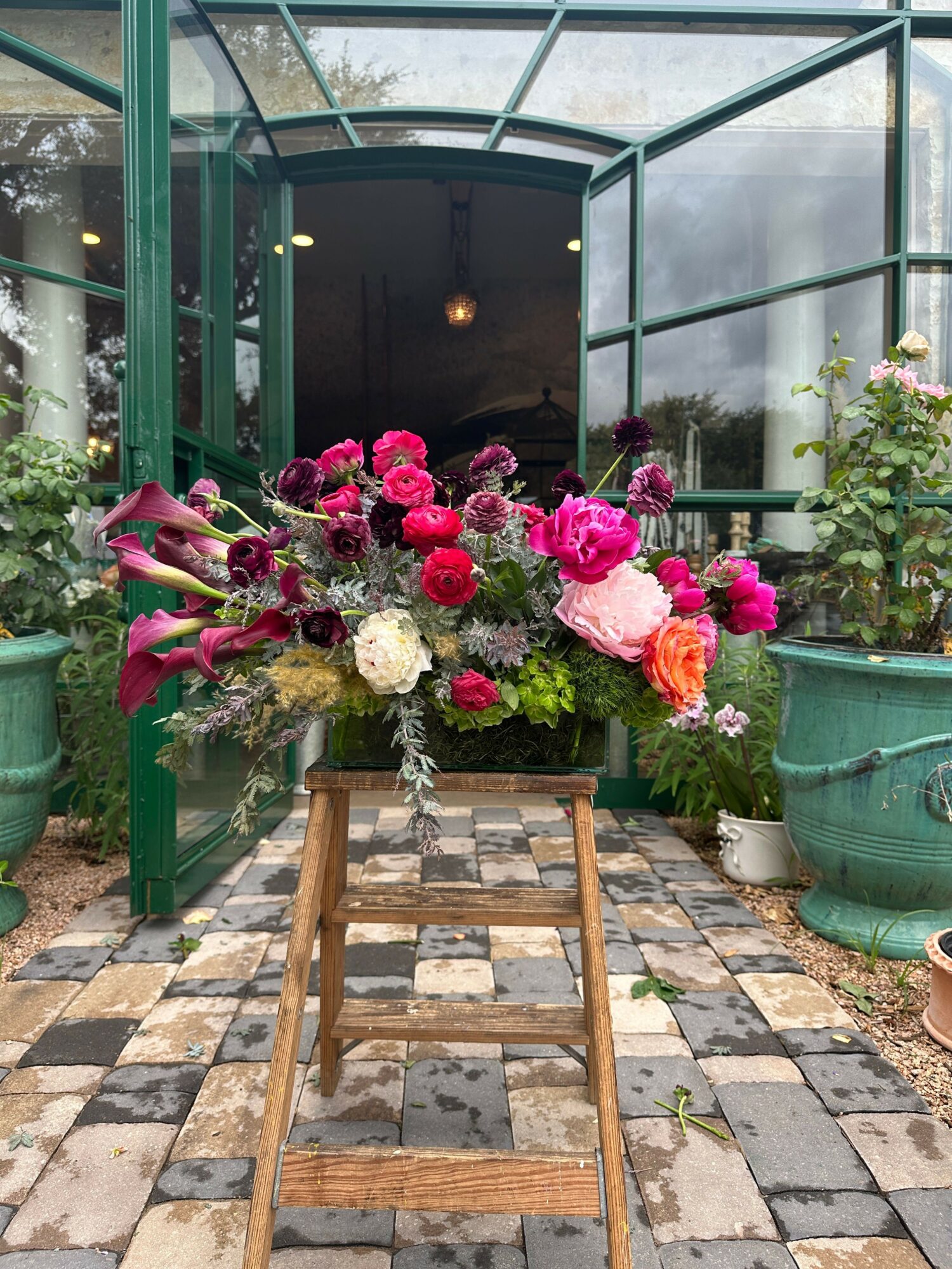 Flower arrangement with pink, white, and purple flowers on a wooden ladder outside a greenhouse.