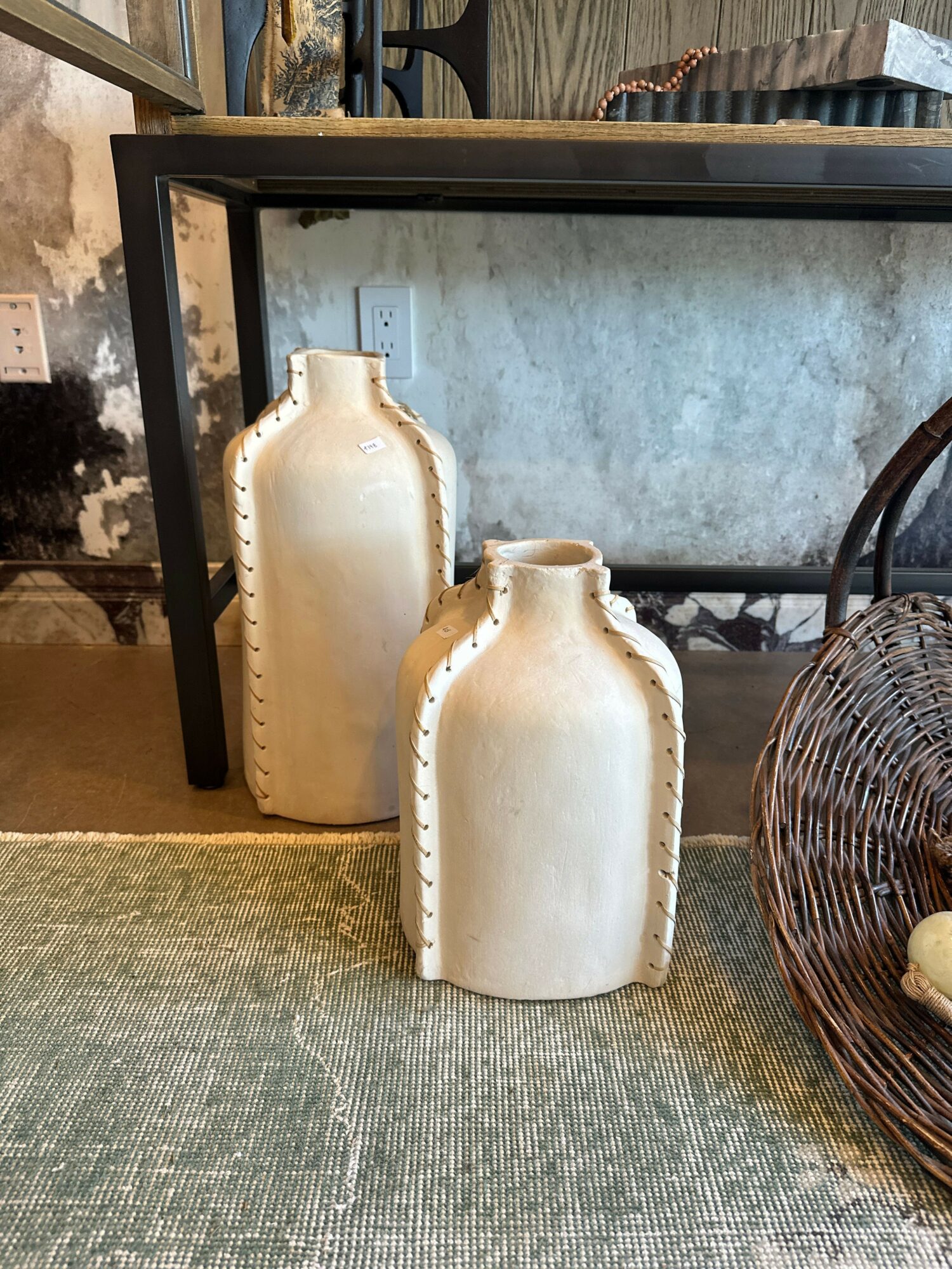 Two cream-colored ceramic vases with decorative edges are placed on a textured surface near a mirror and a wicker basket.